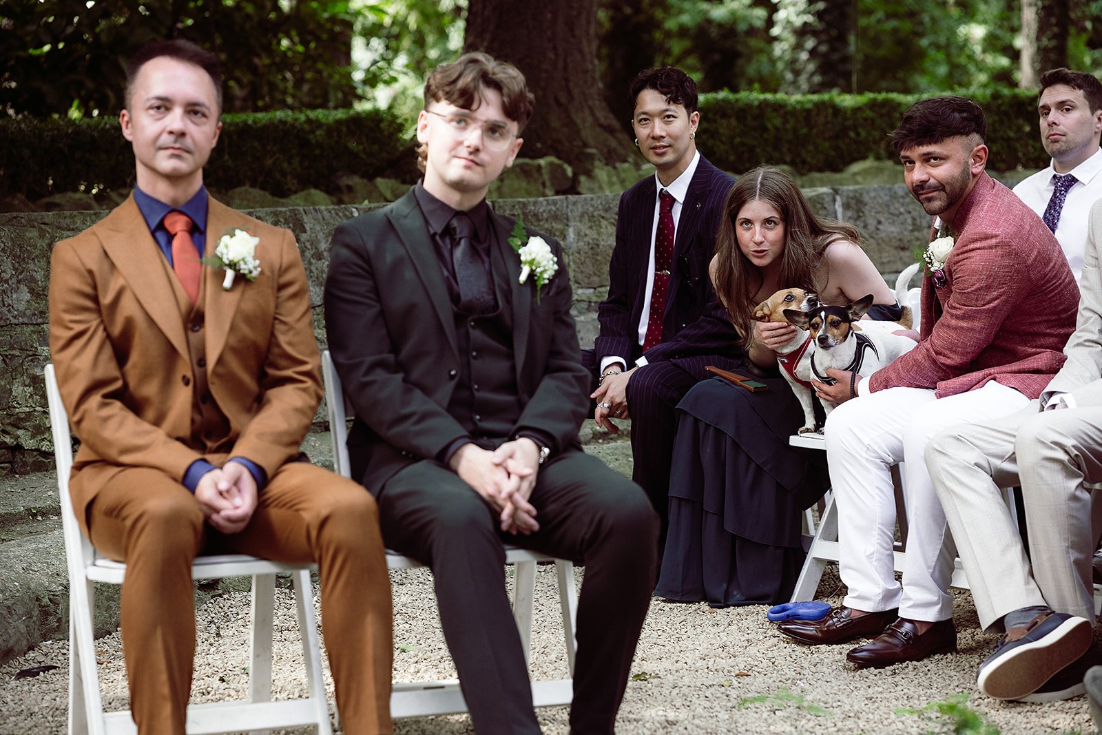 Group of people attending an outdoor wedding ceremony, dressed in formal and semi-formal attire, seated on white chairs with a brick wall and trees in the background. Some individuals are holding dogs.