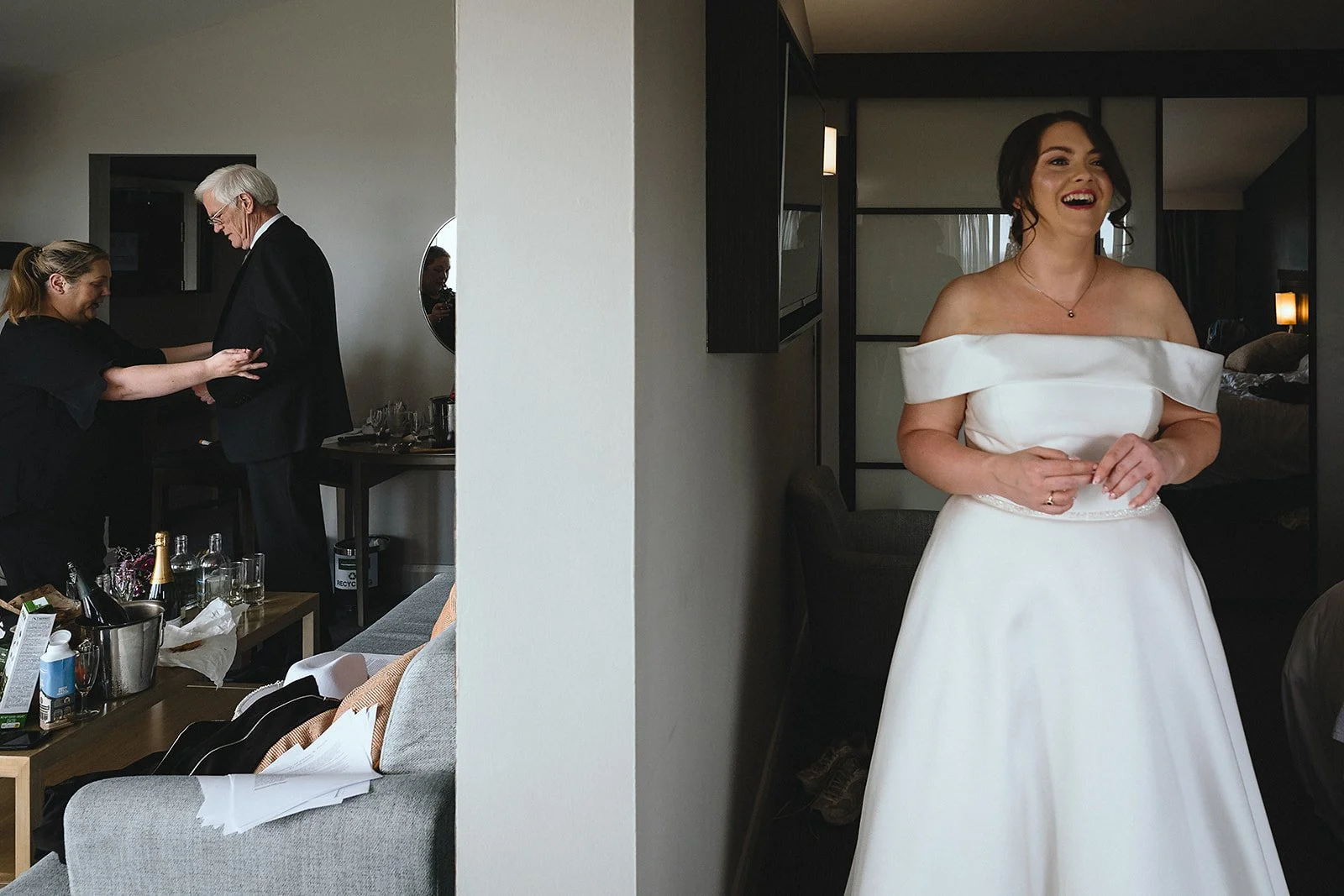 A bride in a white off-the-shoulder wedding dress standing in a hotel room, smiling. On the opposite side, a woman adjusting an older man's suit, with a table of bottles and glassware nearby.