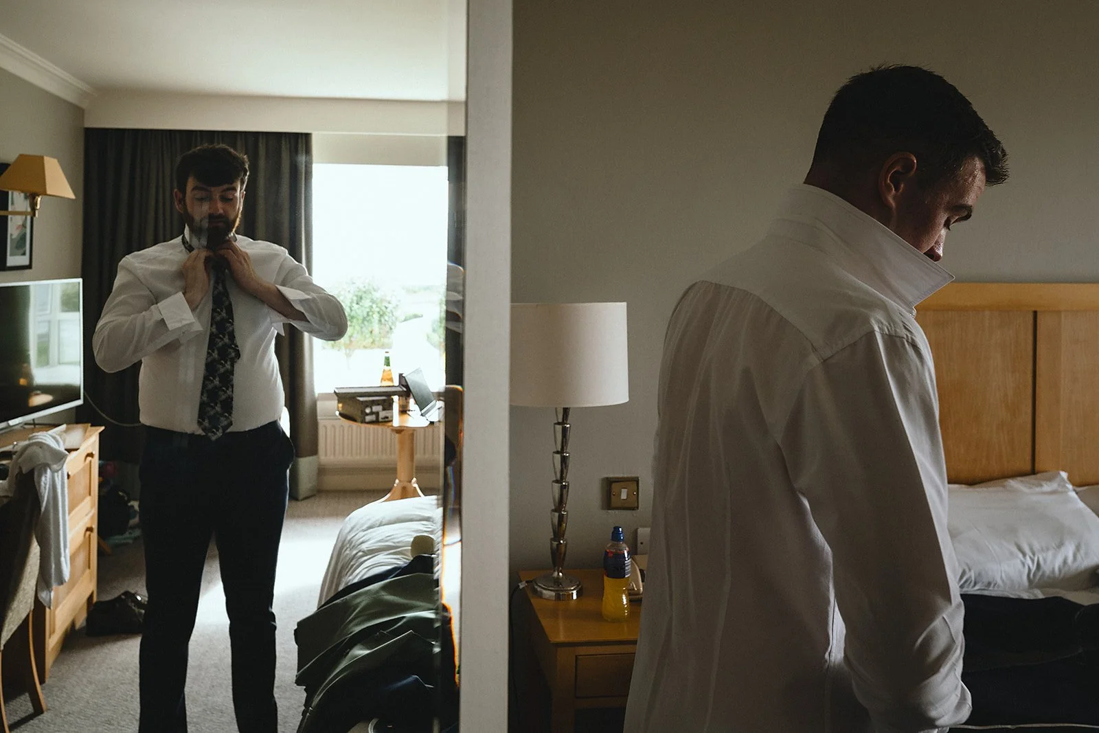 Two men dressing in a hotel room, one is adjusting his tie in front of a mirror, the other is looking down, standing next to a bed.