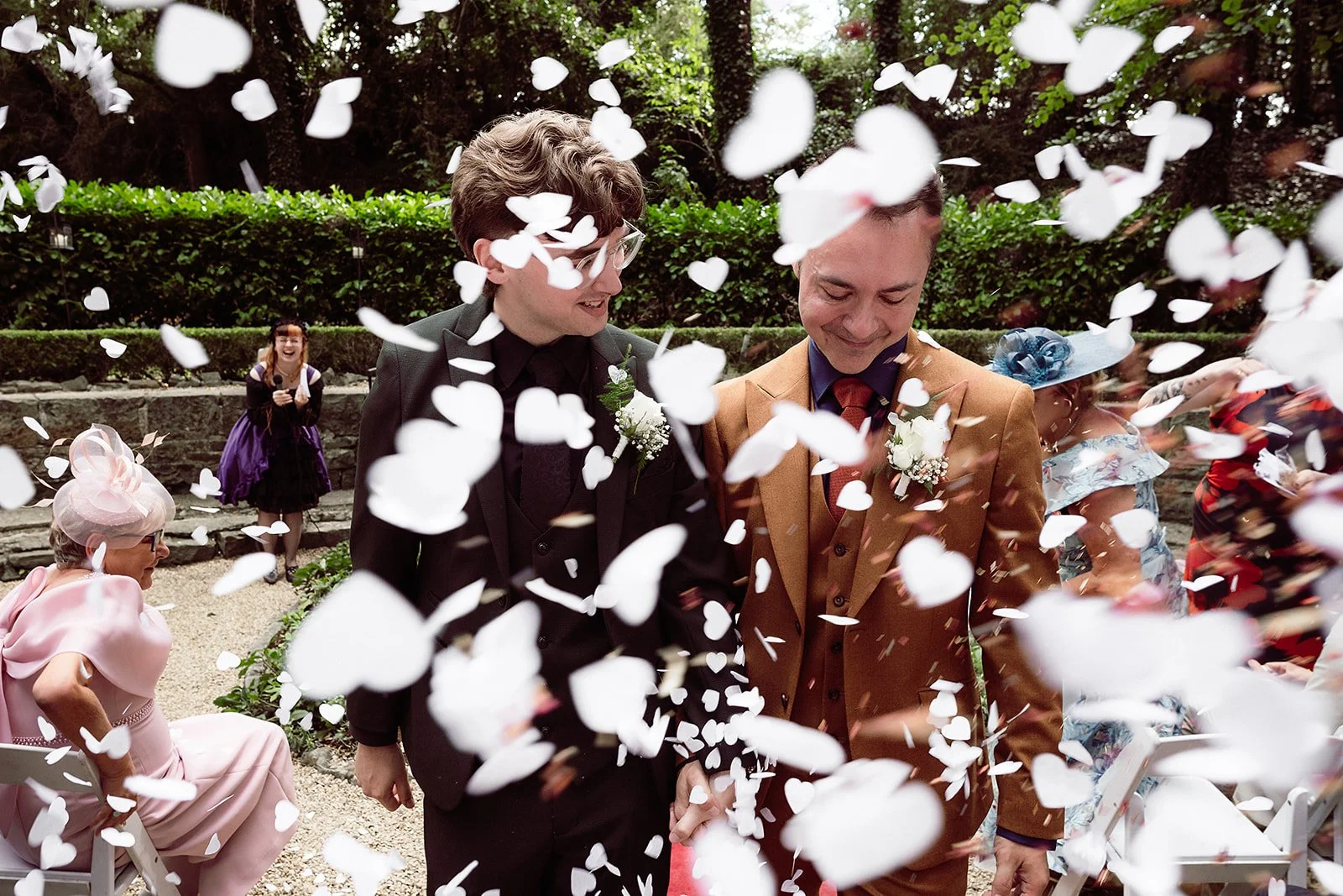 Two men in suits walking hand-in-hand during a wedding celebration with white paper hearts falling around them, others sitting and standing in celebration in the background.