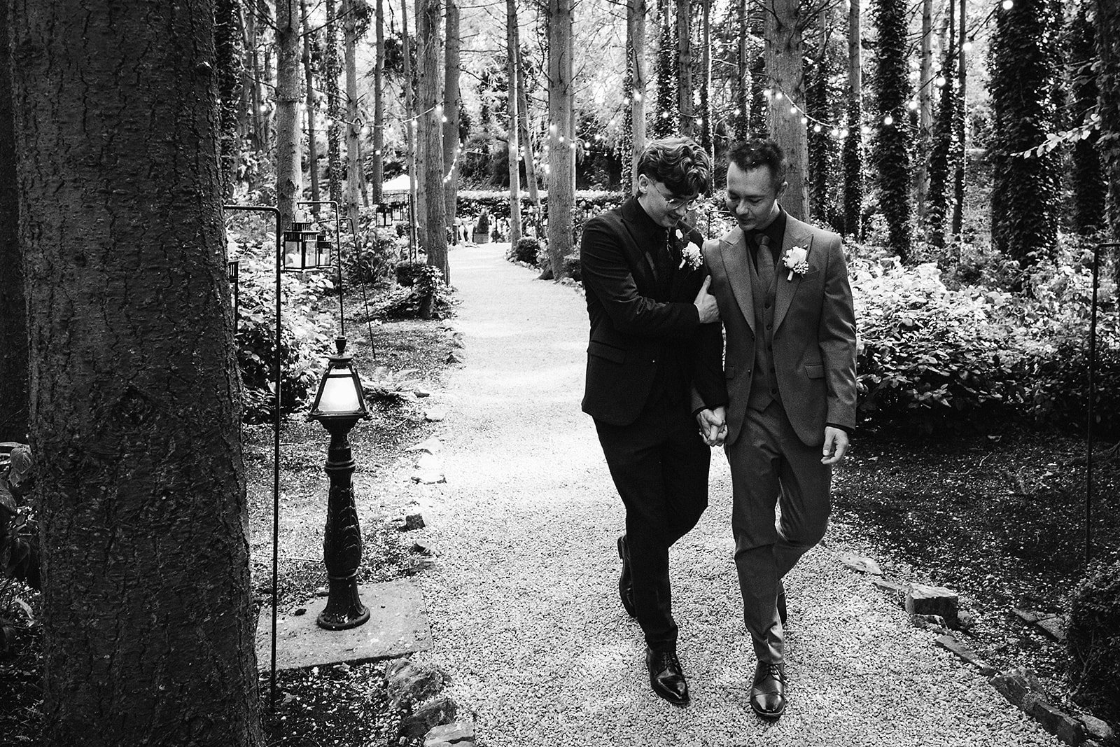 Two men dressed in suits, one in a tuxedo and the other in a blazer, walk hand-in-hand down a wooded path decorated with string lights, holding each other's hands and smiling.