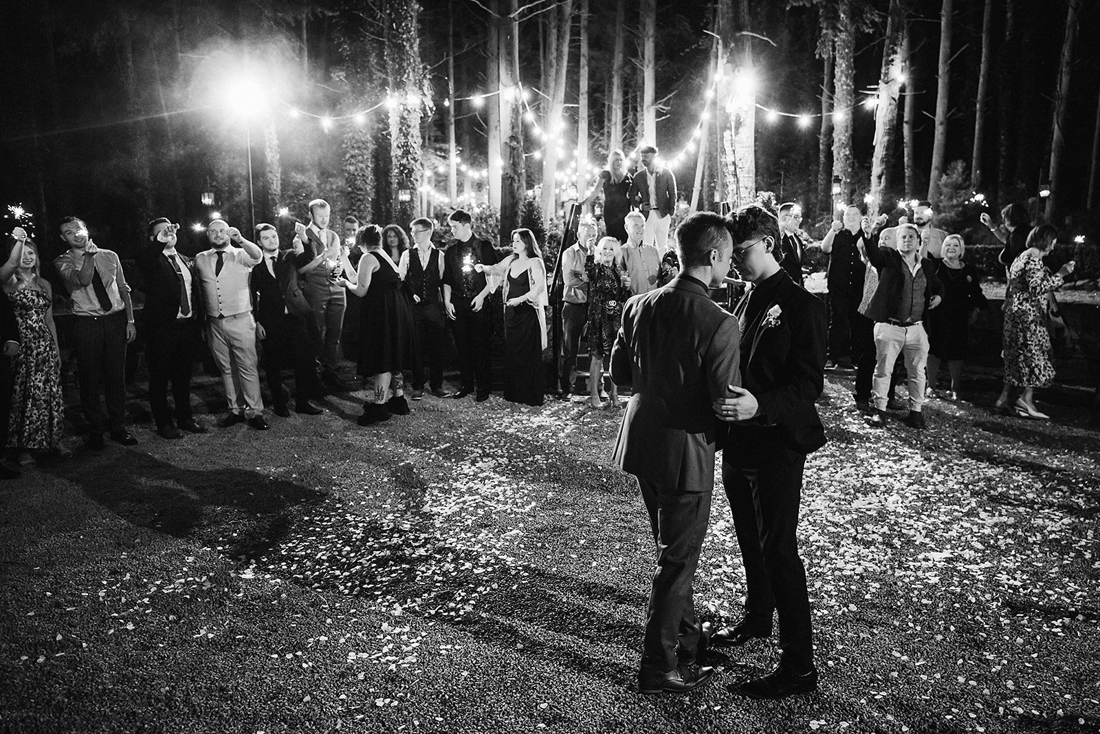 A black and white photo of a nighttime outdoor wedding reception in a wooded area. Two men are dancing or sharing a moment in the foreground, while a large group of people watch and socialize in the background, illuminated by string lights hanging be