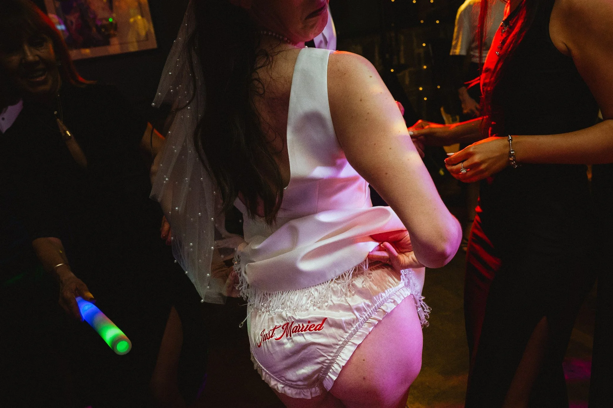 A woman wearing a white satin top and white shorts with the words 'Just Married' embroidered, dancing at a celebration with other women in dark clothing at a party or wedding reception.