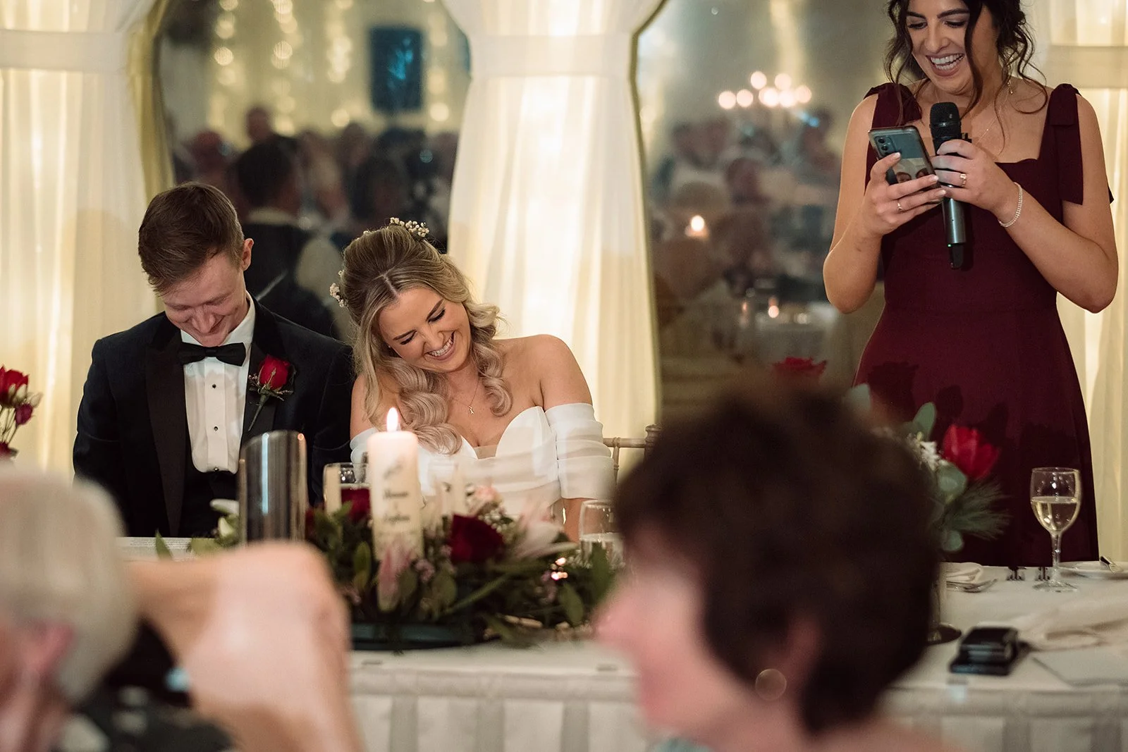 A wedding reception scene with a bride and groom seated at a table, both smiling and leaning towards each other, with a woman in a burgundy dress standing nearby holding a microphone and a phone, possibly giving a speech, in a decorated indoor venue 