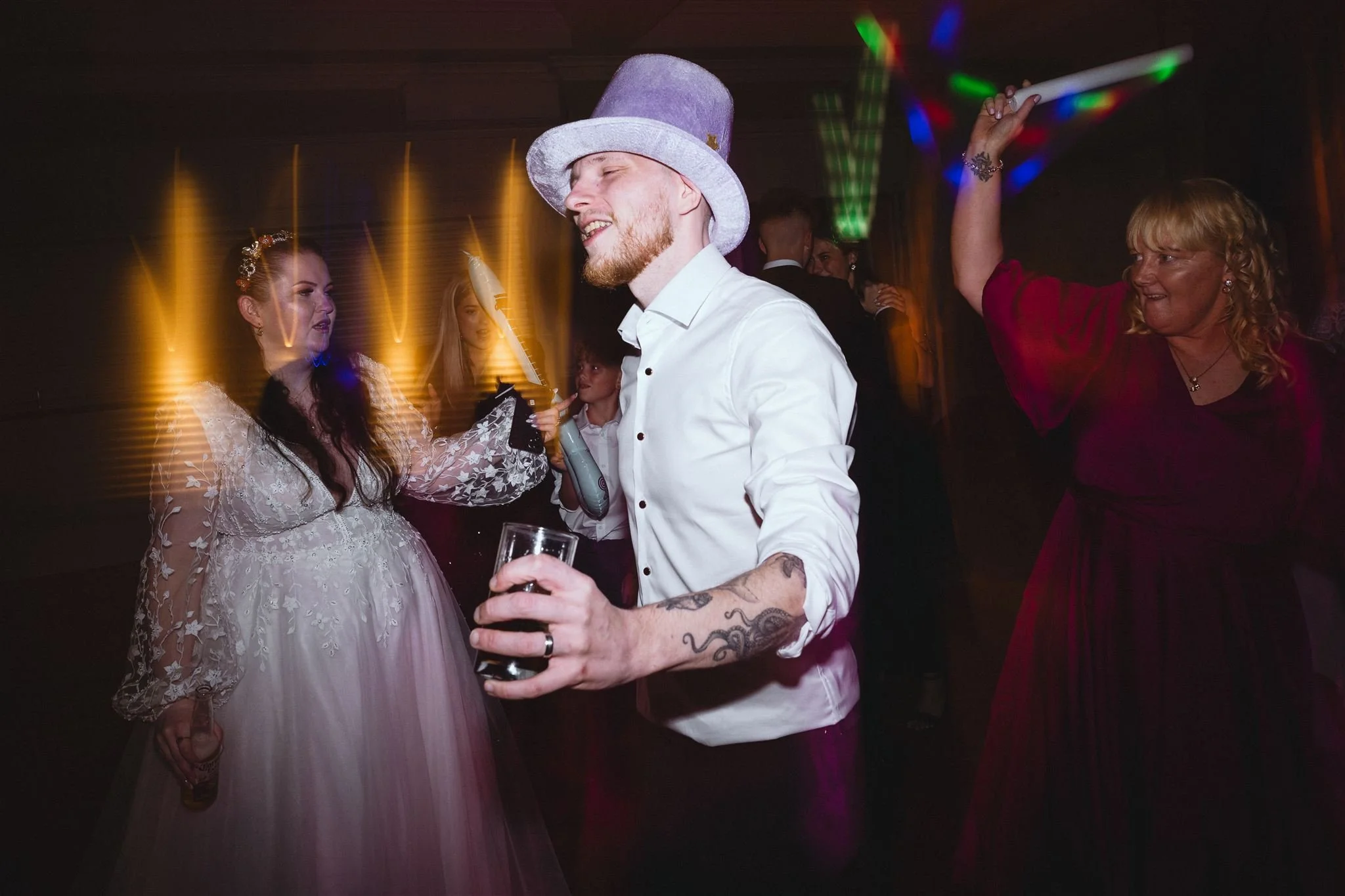 People dancing at a celebration, wearing festive attire, with colorful lights and wooden walls in the background.