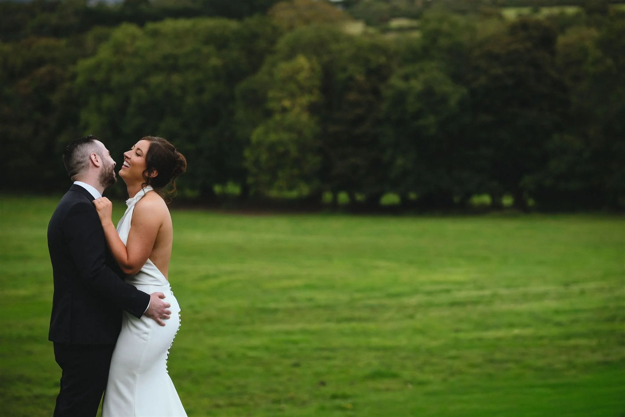A couple dressed in wedding attire, embracing each other in a grassy outdoor setting with trees in the background. The woman is wearing a white wedding gown, and the man is wearing a black suit and white shirt.