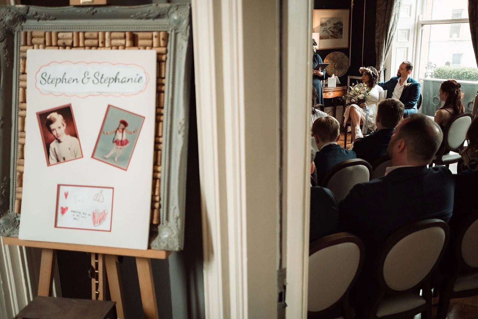 Guests seated in a decorated room listening to a wedding ceremony with a bride and groom sitting together near a window, with a woman officiating.