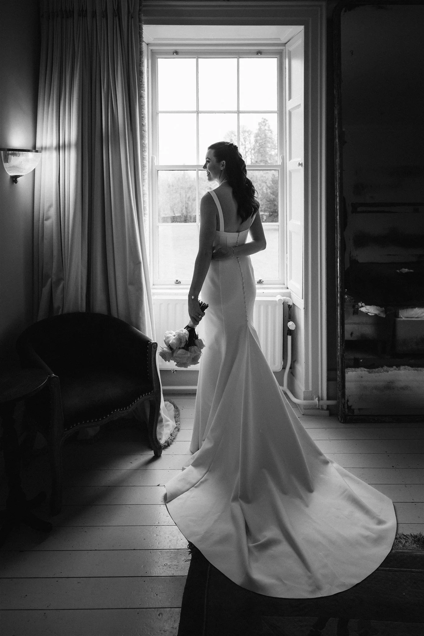 A woman in a wedding dress stands by a large window, holding a bouquet of flowers, with sunlight streaming in from outside.