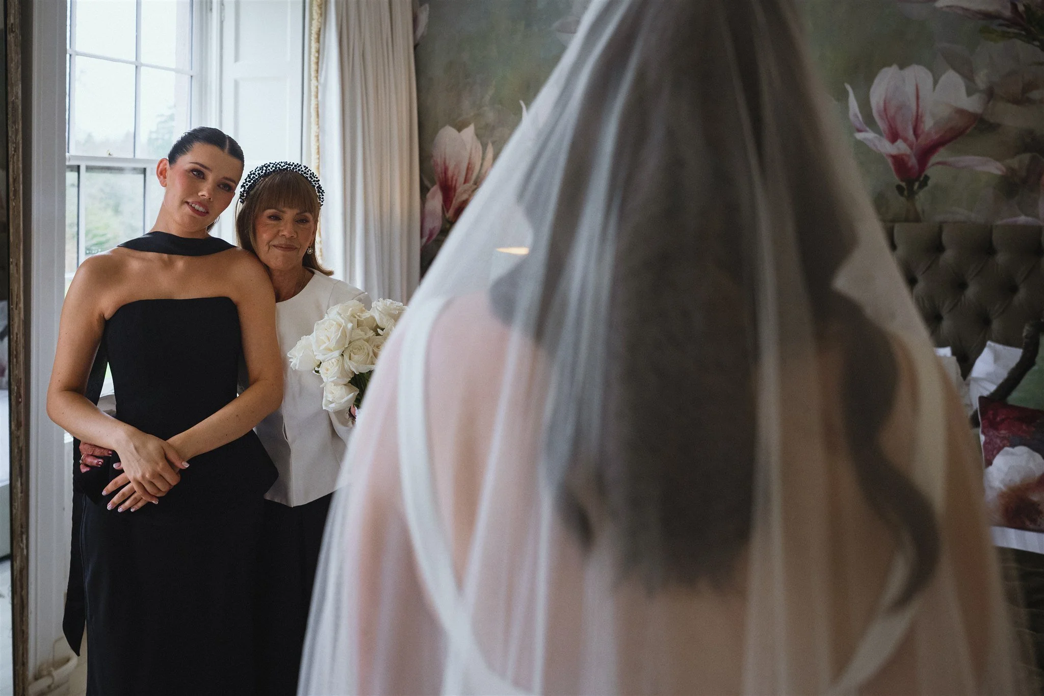 Two women, one younger and one older, standing inside a room with floral wallpaper, smiling at the camera. The younger woman is wearing a black sleeveless dress and the older woman is holding a bouquet of white roses, dressed in white with a headband