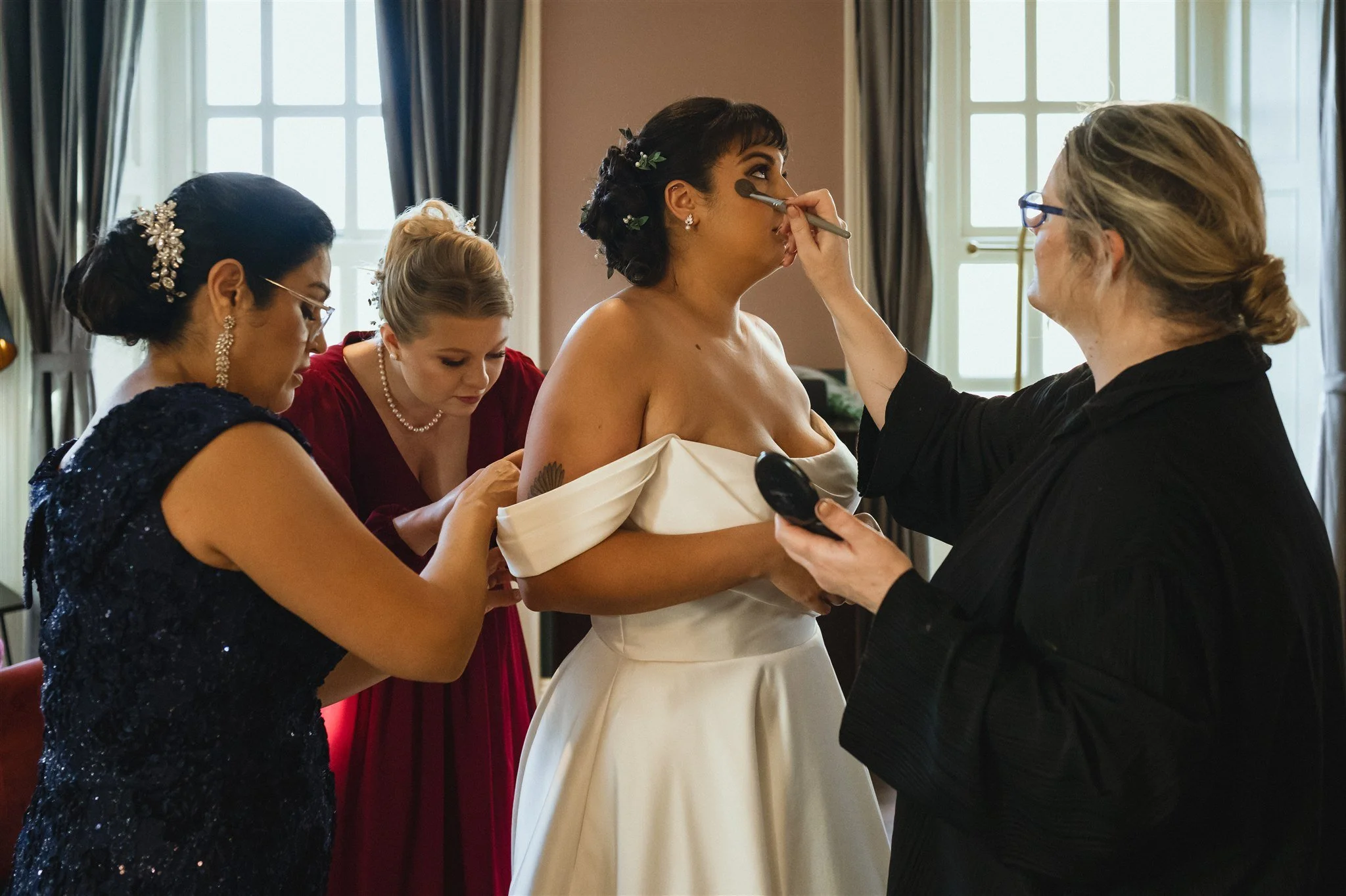 A bride getting her makeup done by a makeup artist while four women look on in a room with large windows and dark curtains.