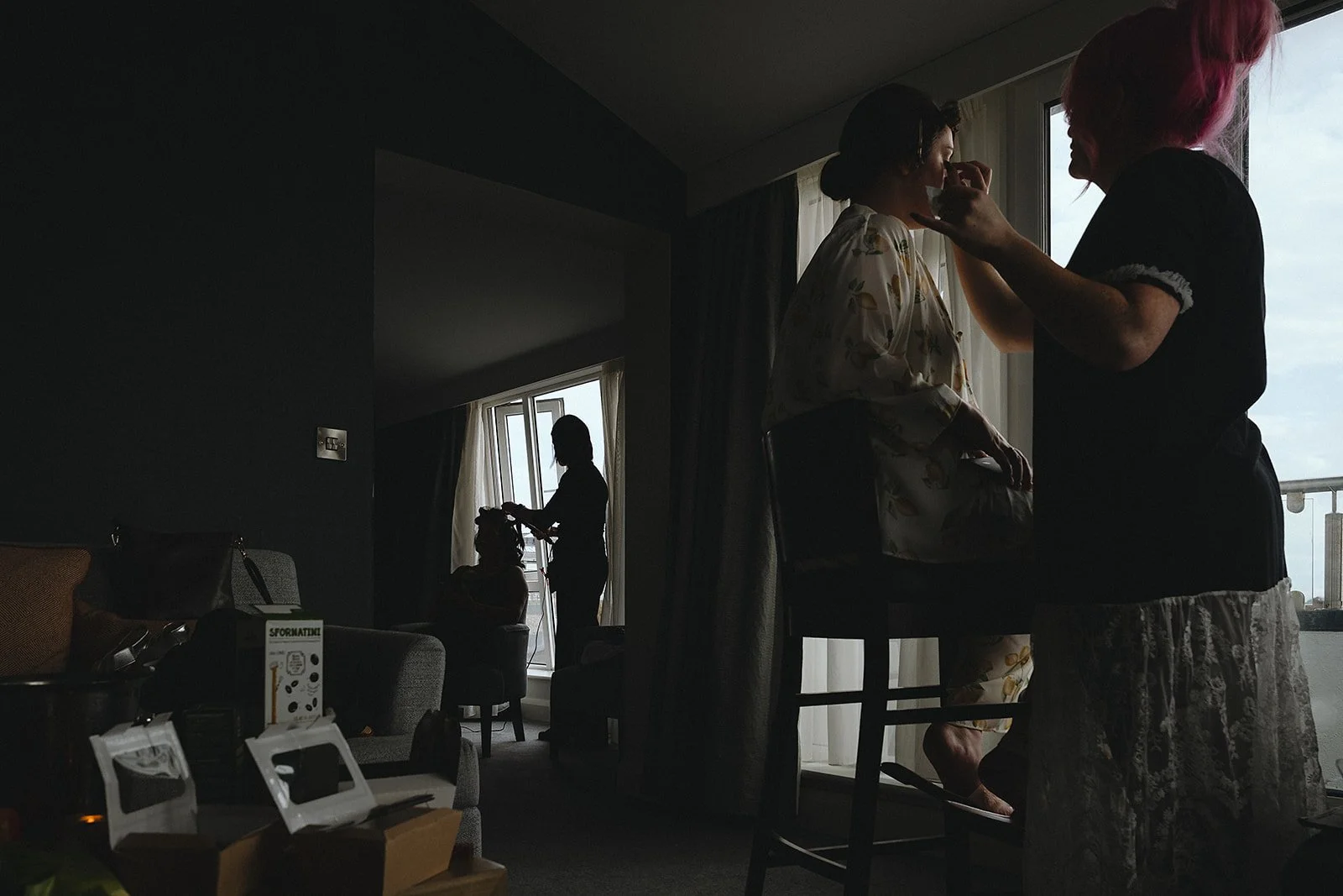 Two women getting ready, one applying makeup to the other, in a dimly lit room with large windows showing a cloudy sky outside.