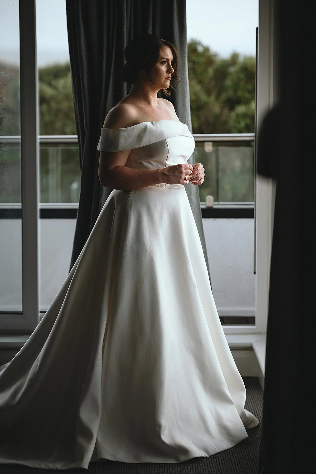 A woman in a white off-the-shoulder wedding gown standing indoors near a large window with curtains, looking contemplative.