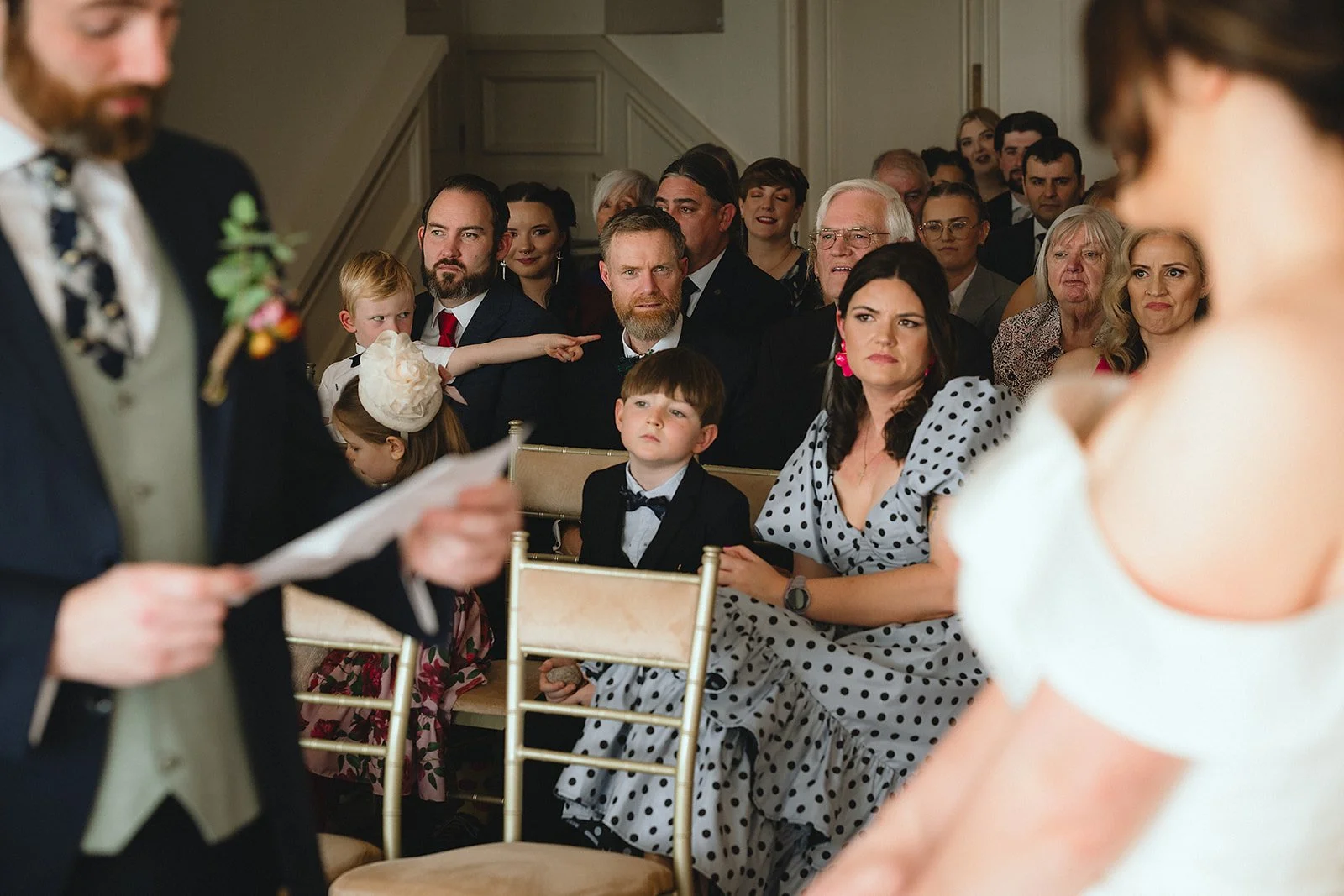 Group of people attending a wedding ceremony, sitting on chairs, facing the bride and groom. Some children and adults look serious or interested, inside a decorated room.