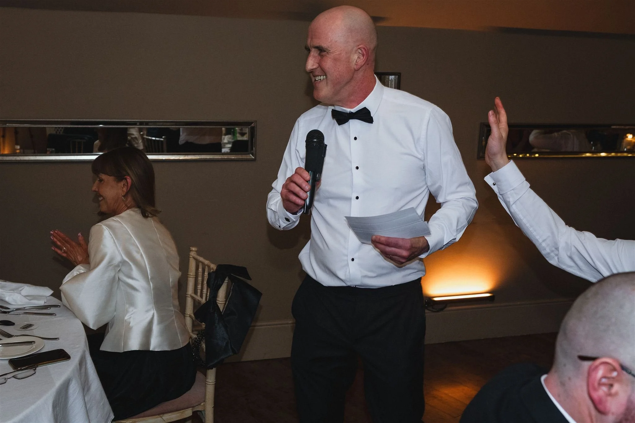 A man in a white dress shirt and black bowtie giving a speech with a microphone at a formal event, while women sit at a table clapping and smiling.