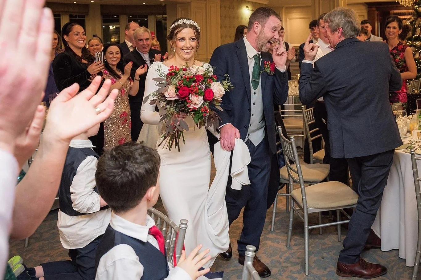 Bride holding a bouquet and smiling as she walks through a wedding reception surrounded by guests, celebrating.