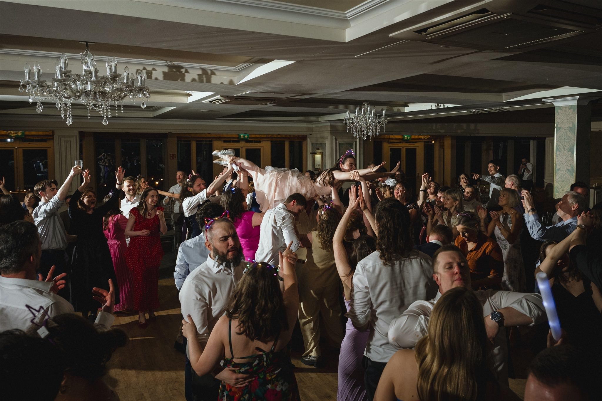 People at a party or celebration, with a woman in a pink dress being lifted on others' hands in a large room with chandeliers and windows.
