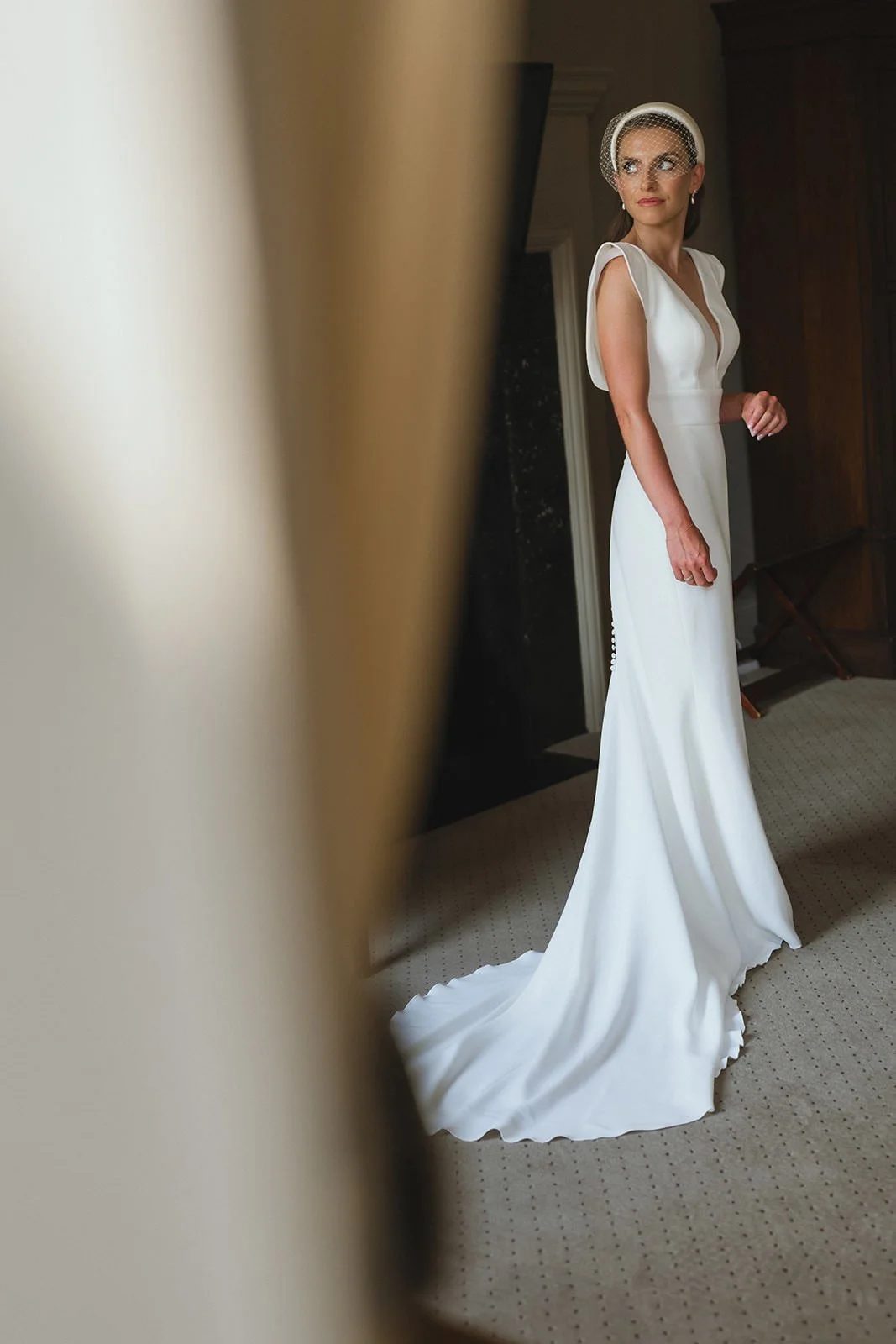 A woman in a white wedding dress with a headpiece and net veil standing in a room.