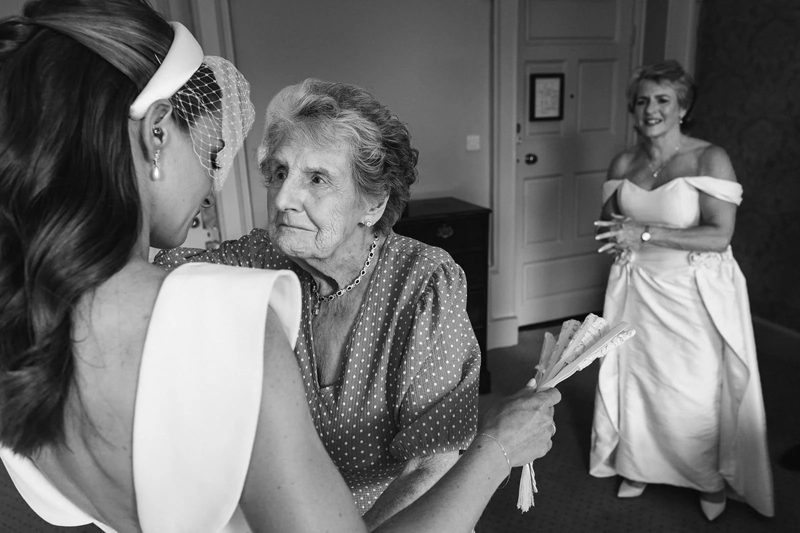 A bride with long dark hair and pearl earrings is face to face with an elderly woman, who is holding a fan. A woman in a white off-shoulder wedding dress stands in the background, smiling, in a decorated room.