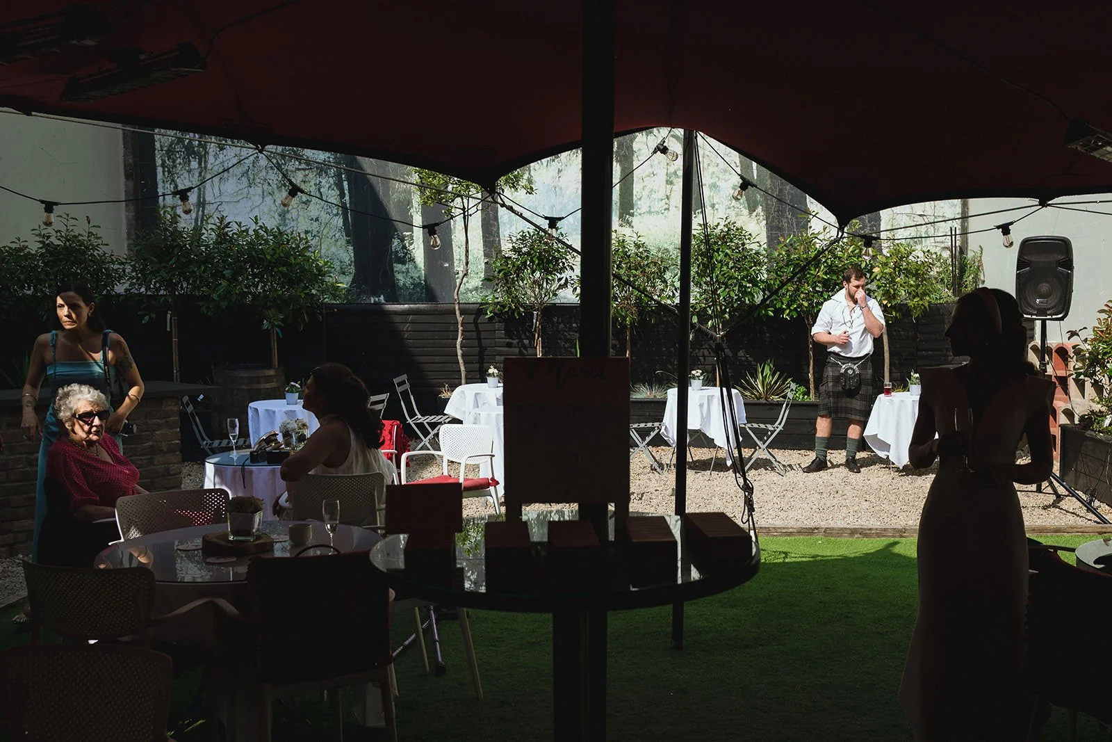 Outdoor restaurant or event space with tables, chairs, and string lights, featuring people seated at tables and a performer in Scottish attire on stage.