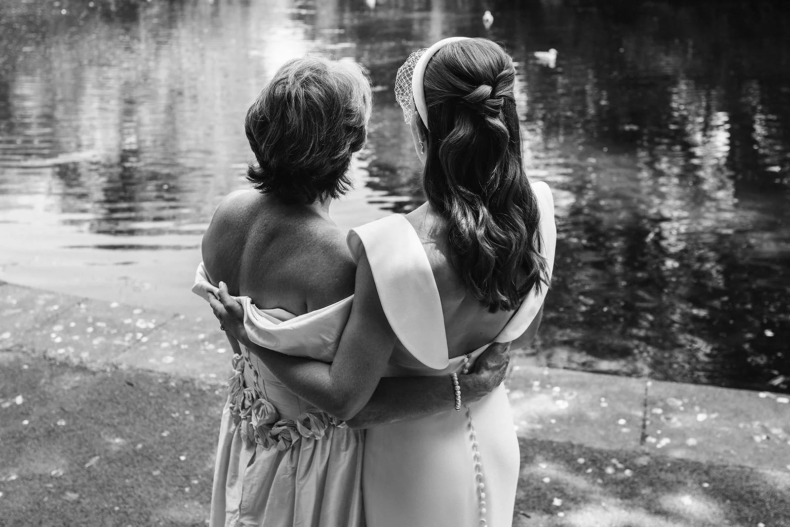 Black and white photo of two women embracing by a body of water, one appears older and the other younger, both wearing elegant dresses, one has a headband and a veil, and the scene seems emotionally tender.