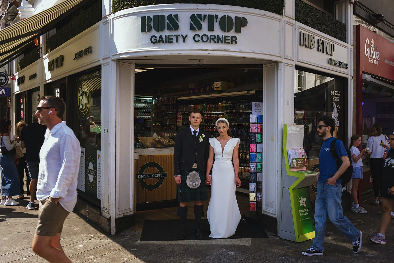 A bride and groom in wedding attire standing in front of a small coffee shop called 'Bus Stop Gaiety Corner' in an urban setting, surrounded by pedestrians.