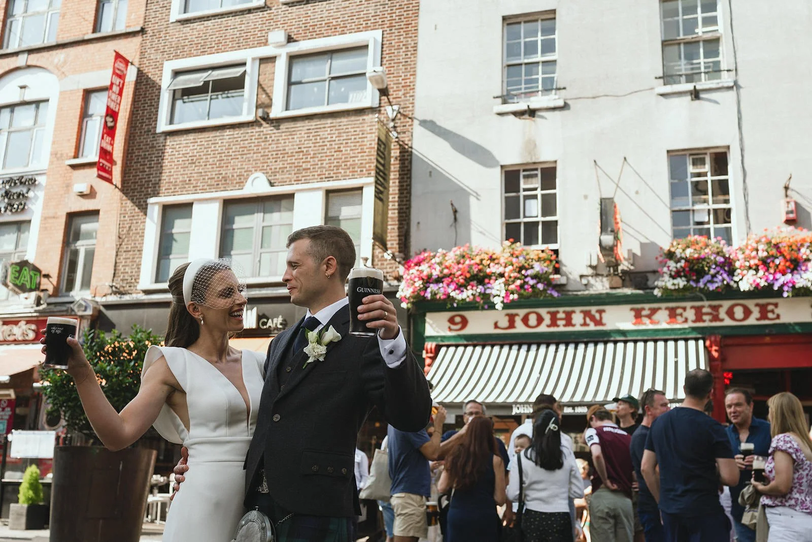 A bride and groom smiling and holding Guinness beer pints in a busy city street during daytime. The bride wears a white dress with a birdcage veil, and the groom is in a black suit with a white shirt and boutonniere. People are gathered around, and c