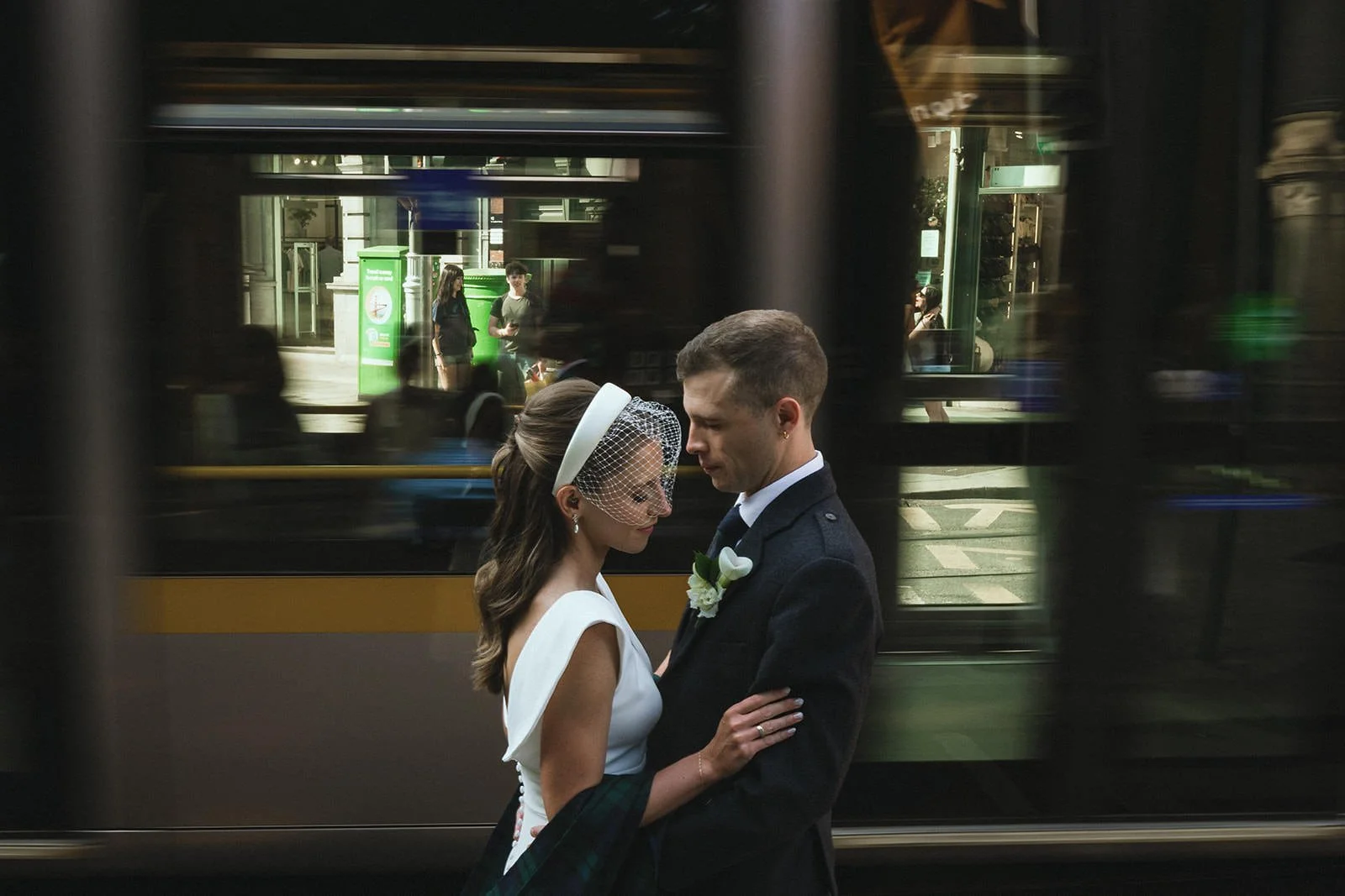 A bride and groom sharing a moment, standing closely with eyes closed, on the street in front of a passing tram, with city buildings and pedestrians in the background.