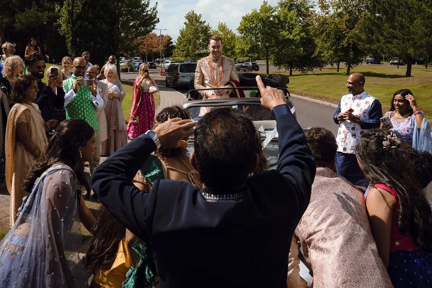 A wedding celebration outdoors with a diverse group of people dressed in colorful traditional attire, surrounding a decorated vehicle with the groom standing on top, smiling and holding a document, while guests take photos and videos.