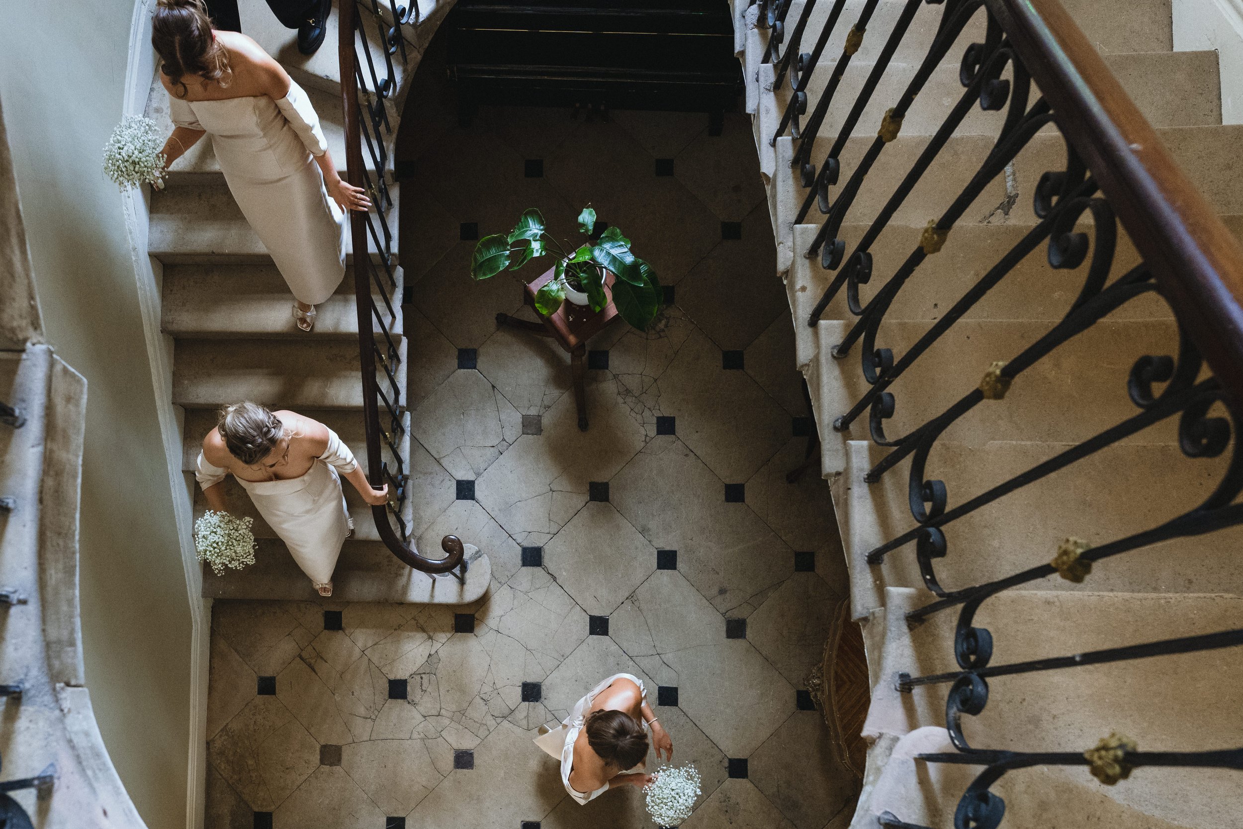 From an overhead view, three women in white dresses are descending a staircase while holding small bouquets of white flowers, with a green potted plant on a small table at the bottom of the stairs.