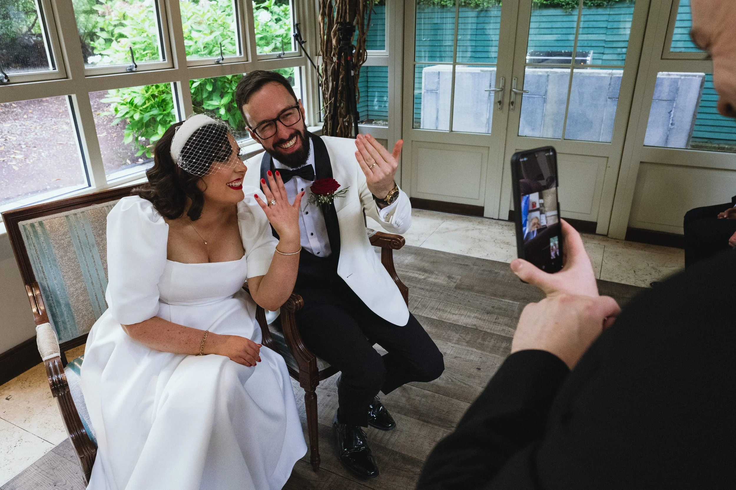 A bride and groom sitting on a vintage wooden bench, smiling and showing their wedding rings, as a person takes a photo of them with a smartphone. The bride wears a white dress and birdcage veil, and the groom wears a tuxedo with a bow tie and boutonniere. They are indoors with large windows and greenery outside.
