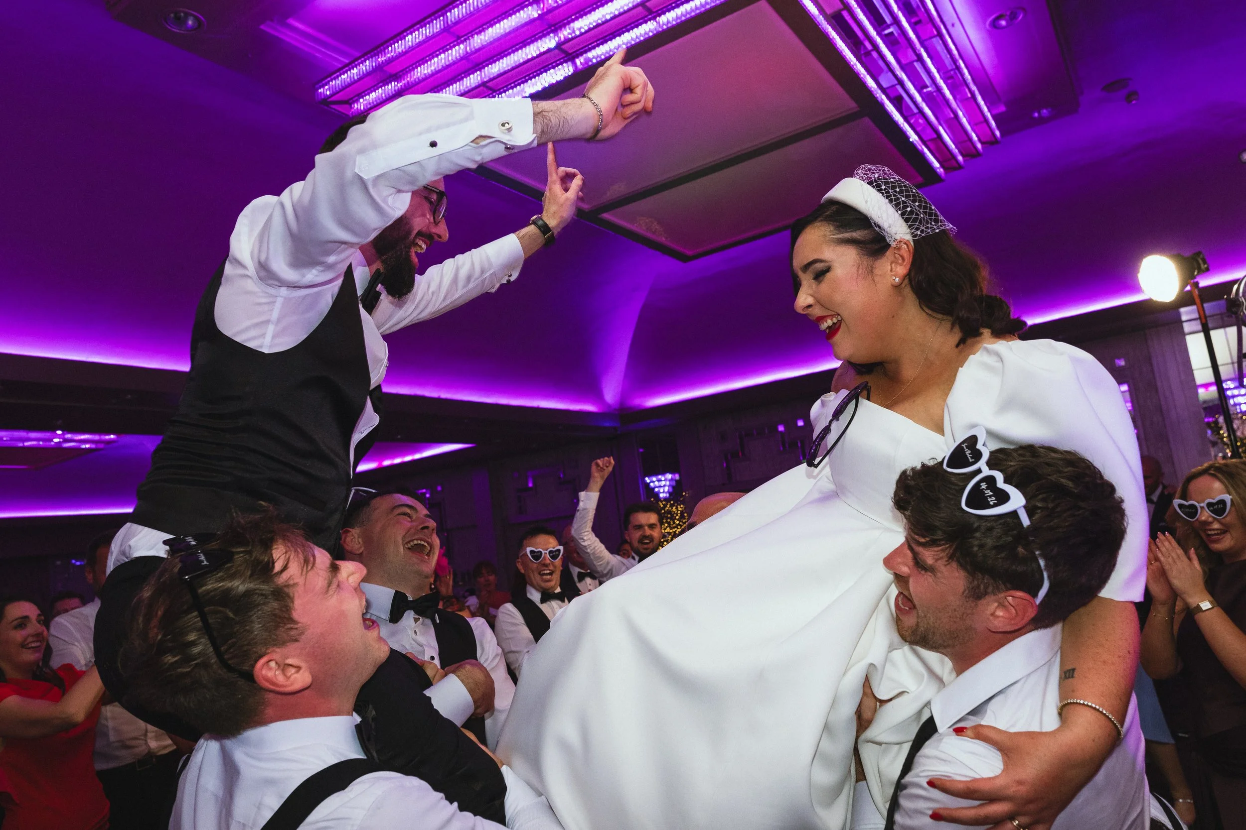 People celebrating at a wedding reception, with the bride being lifted by friends, and the atmosphere illuminated with purple lighting.