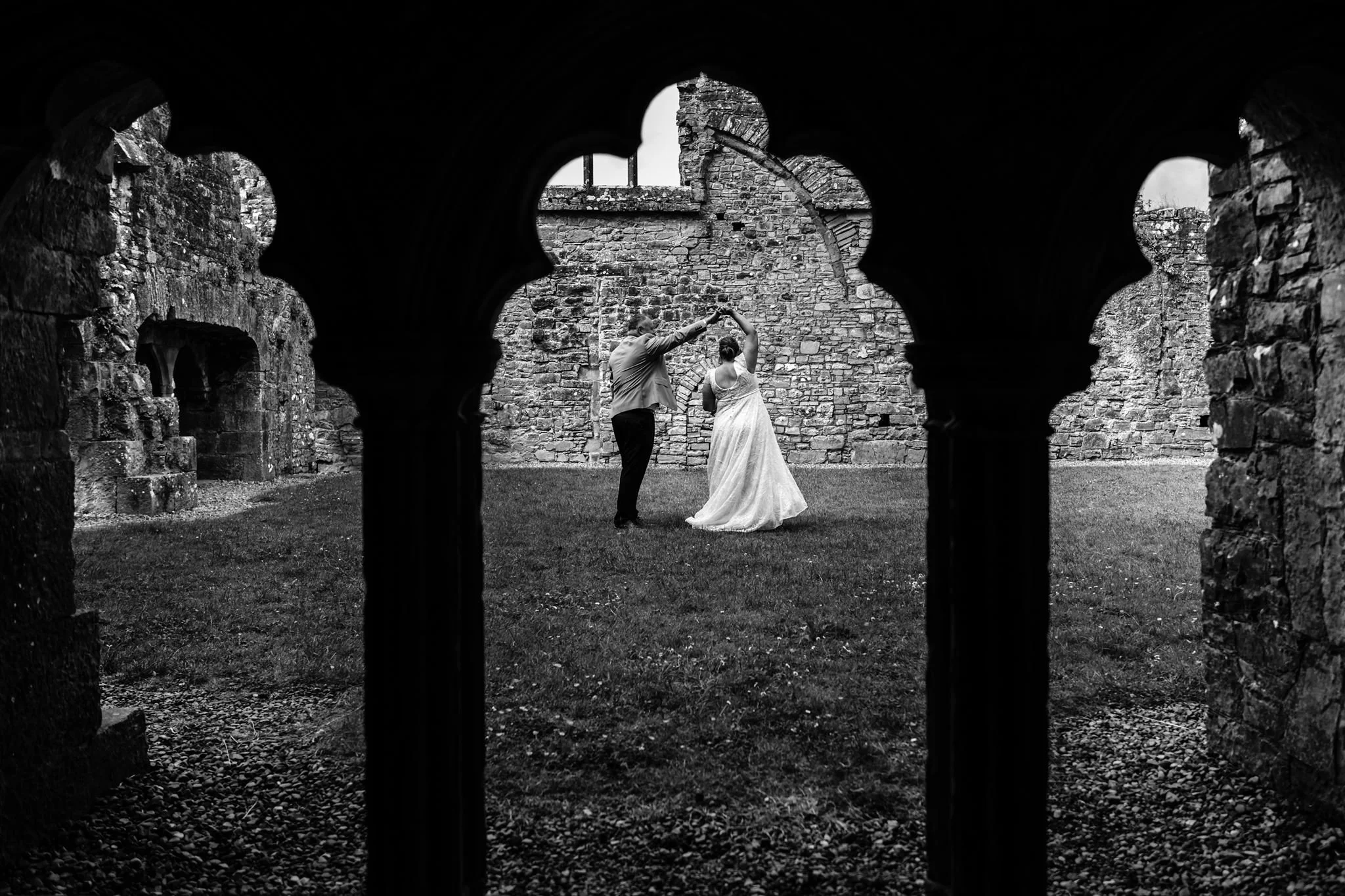 Black and white photo of a couple dancing in a historic courtyard, framed by stone archways.