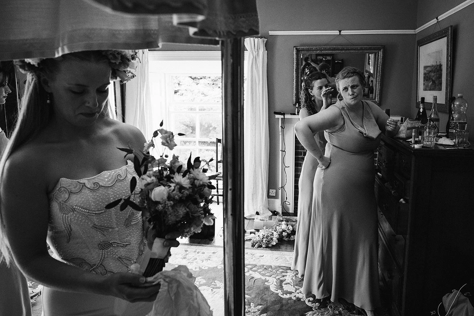 A bride with a floral crown and her bridesmaids in a room, one holding a bouquet, as they prepare for a wedding.