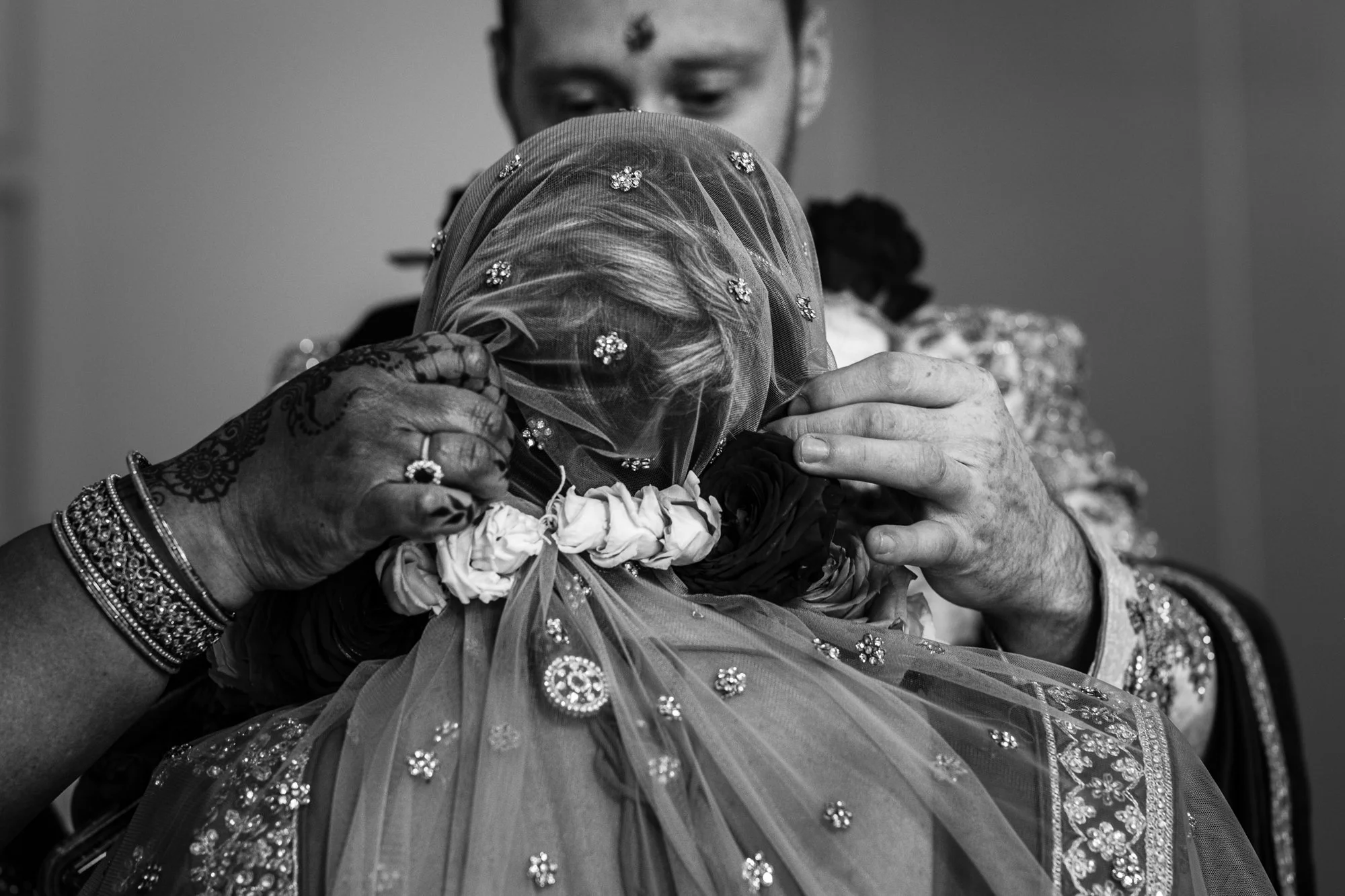 A person adjusting a bride's veil adorned with floral embroidery, with henna-decorated hands and floral garland, during a wedding ceremony.