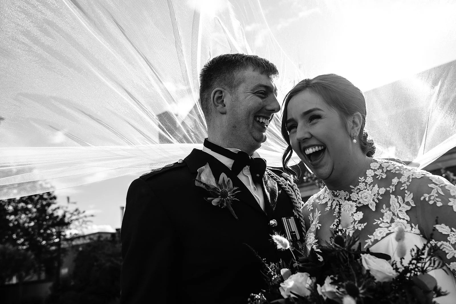 Black and white photo of a joyful bride and groom under a veil. The bride is wearing a lace wedding dress and holding a bouquet, while the groom is in a formal suit with military decorations. They are laughing together.