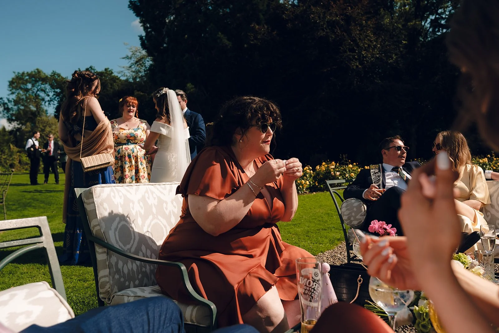 People at an outdoor wedding reception, some sitting on chairs and others standing and conversing, with lush green grass and trees in the background under a bright blue sky.
