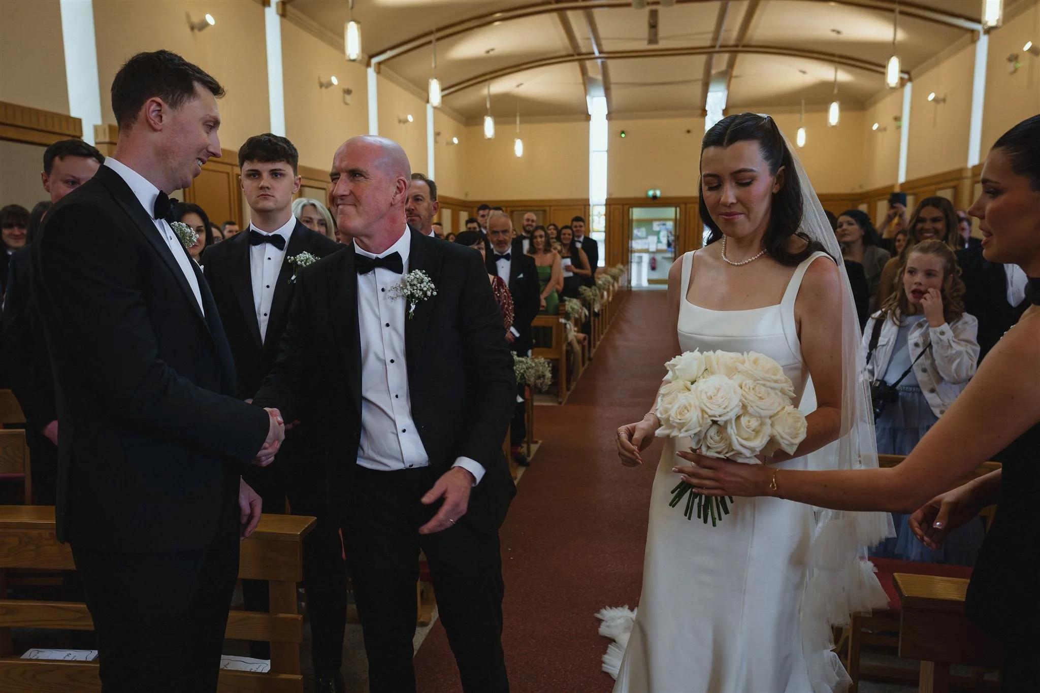 A bride in a white wedding gown holding a bouquet of white roses, standing in a church during a wedding ceremony, with guests seated in the background.