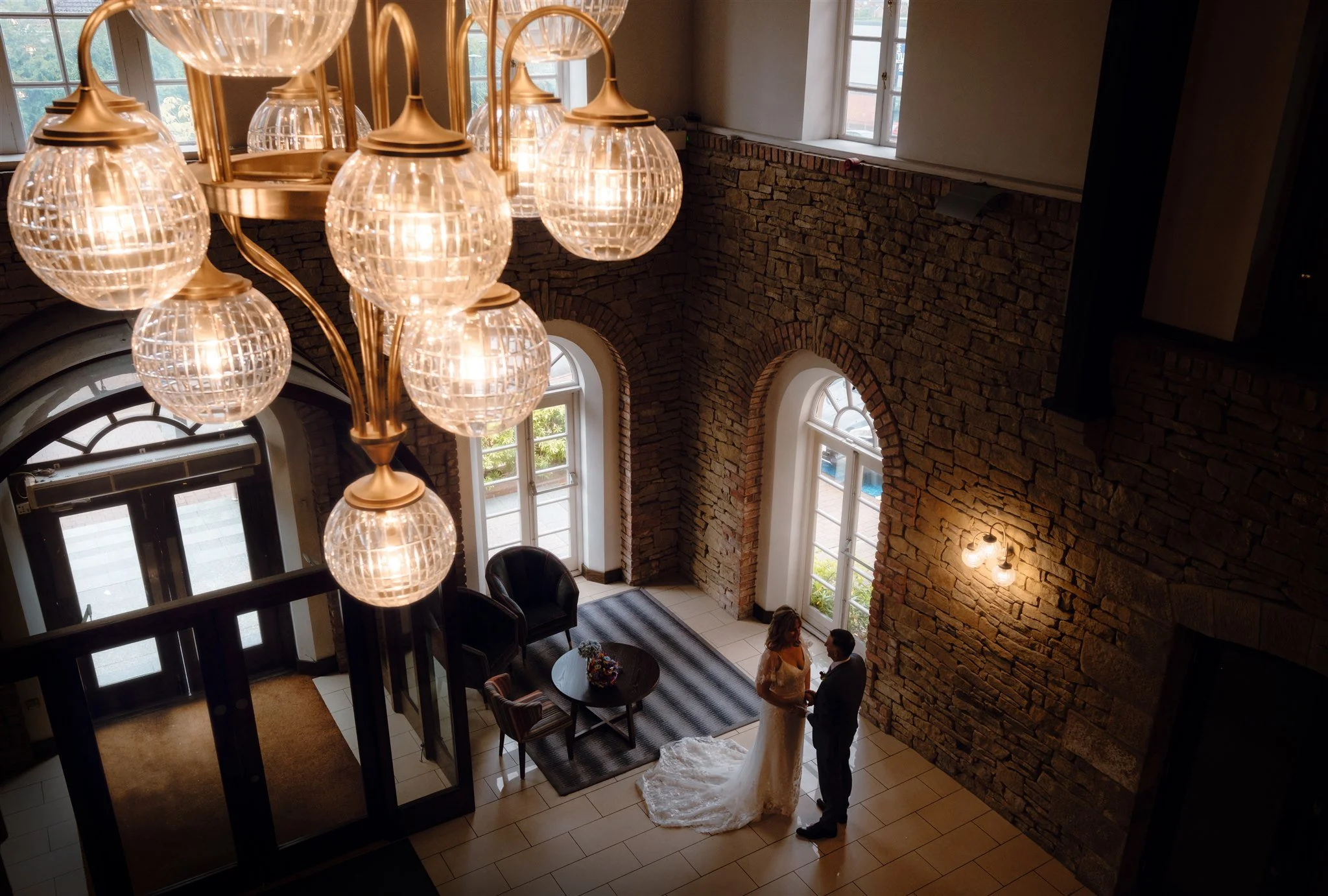 Interior view of a building with a brick wall, large windows, and a chandelier with multiple glass globes hanging from the ceiling. There are two women talking near the entrance area, which has chairs and a small table with flowers.