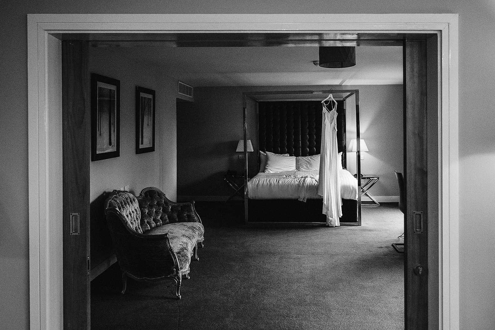 A hotel room with a canopy bed, a swinging wedding dress, a vintage sofa, a lamp on a side table, and framed artwork on the wall, all in black and white.