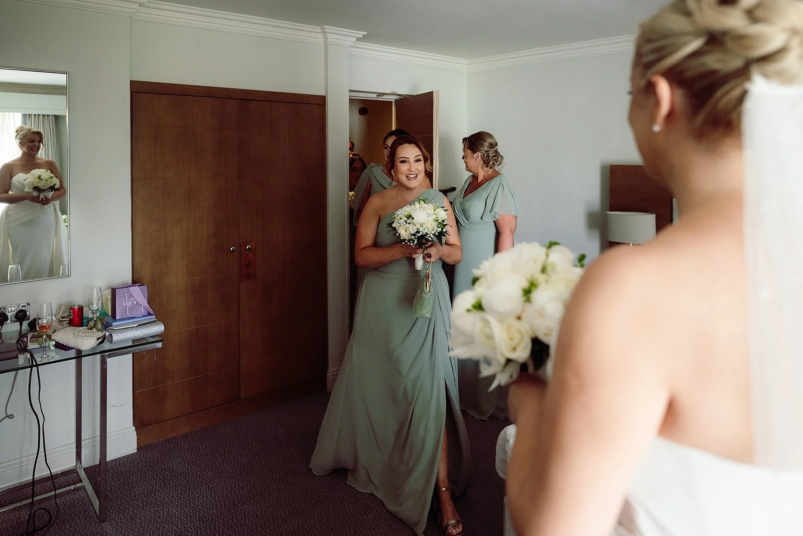 A bride holding a bouquet of white flowers in the foreground, with a woman in a grey dress holding a similar bouquet walking towards her in a hotel room.