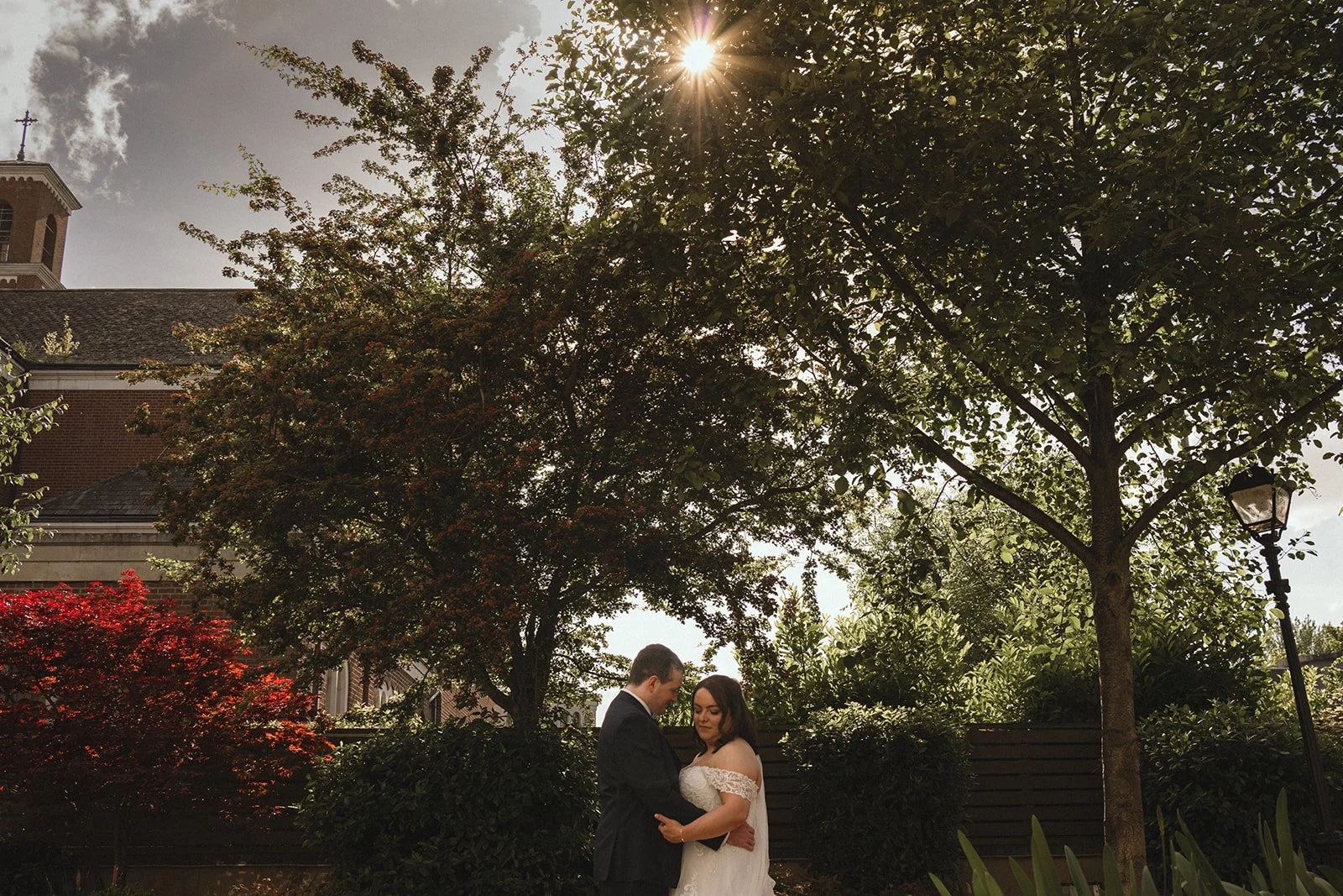 A couple dressed in wedding attire standing outdoors under a large tree with sunlight shining through the branches, surrounded by greenery and a red bush.