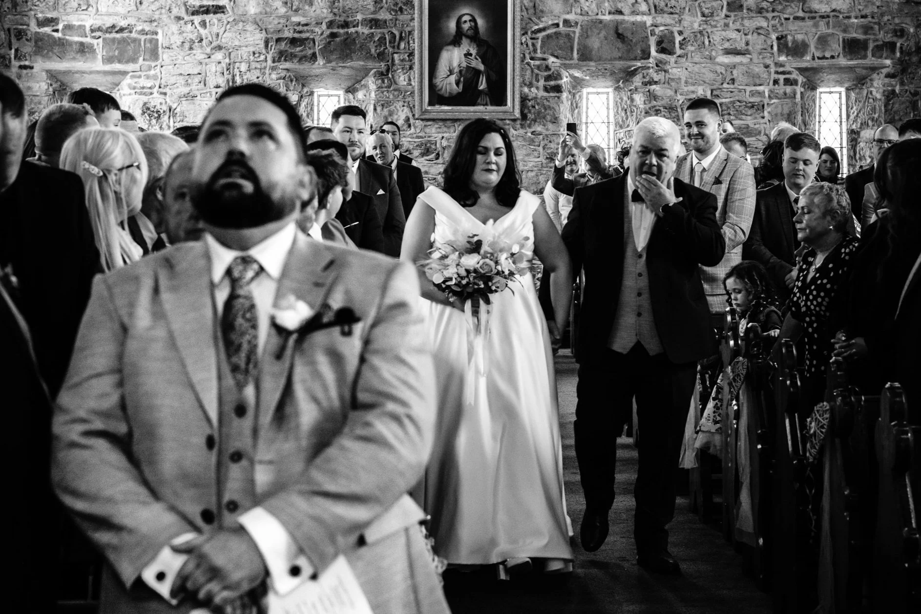 Bride walking down the aisle with a bouquet, accompanied by a person, in a church filled with guests.