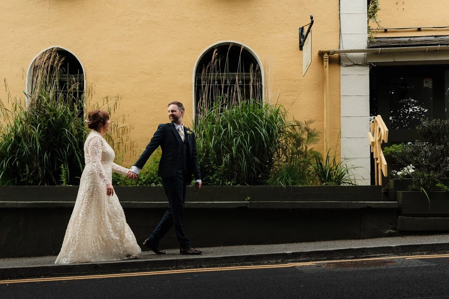A bride in a lace wedding dress and a groom in a dark suit holding hands and walking along a sidewalk, smiling at each other, with a yellow building and green plants in the background.