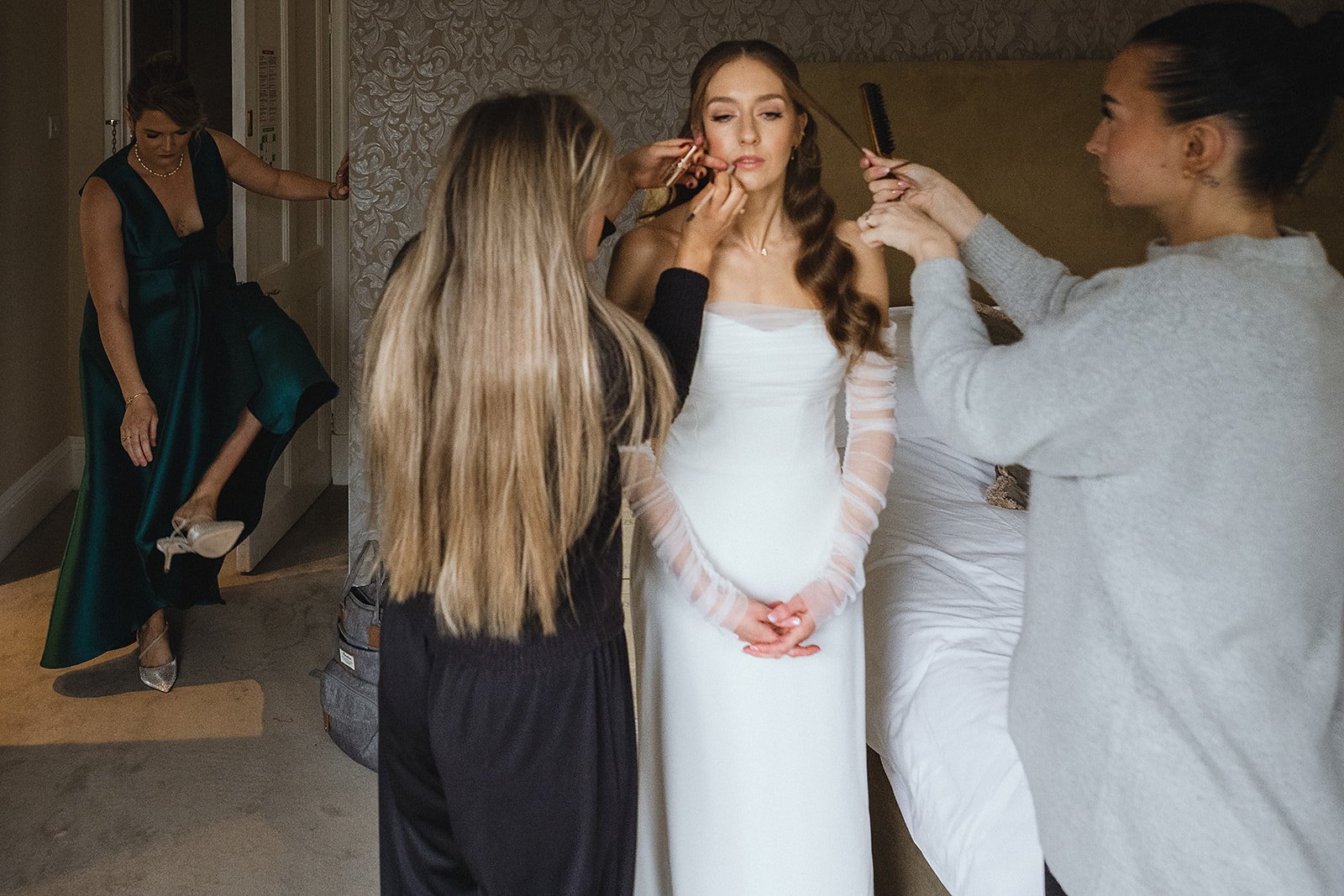 Women getting makeup and hair done before a wedding, with a woman in a white wedding dress in the center.