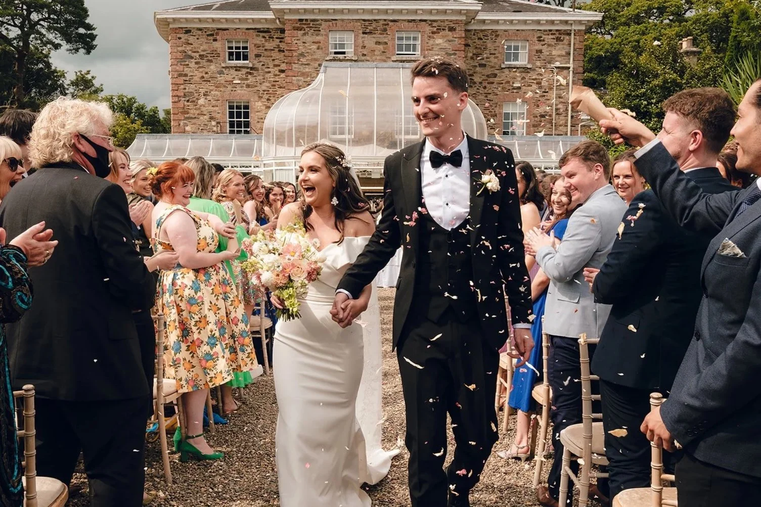 A newlywed couple is walking down the aisle hand in hand, surrounded by smiling guests at an outdoor wedding celebration. Confetti is falling around them, and the bride is holding a bouquet of flowers while everyone is joyful and clapping.