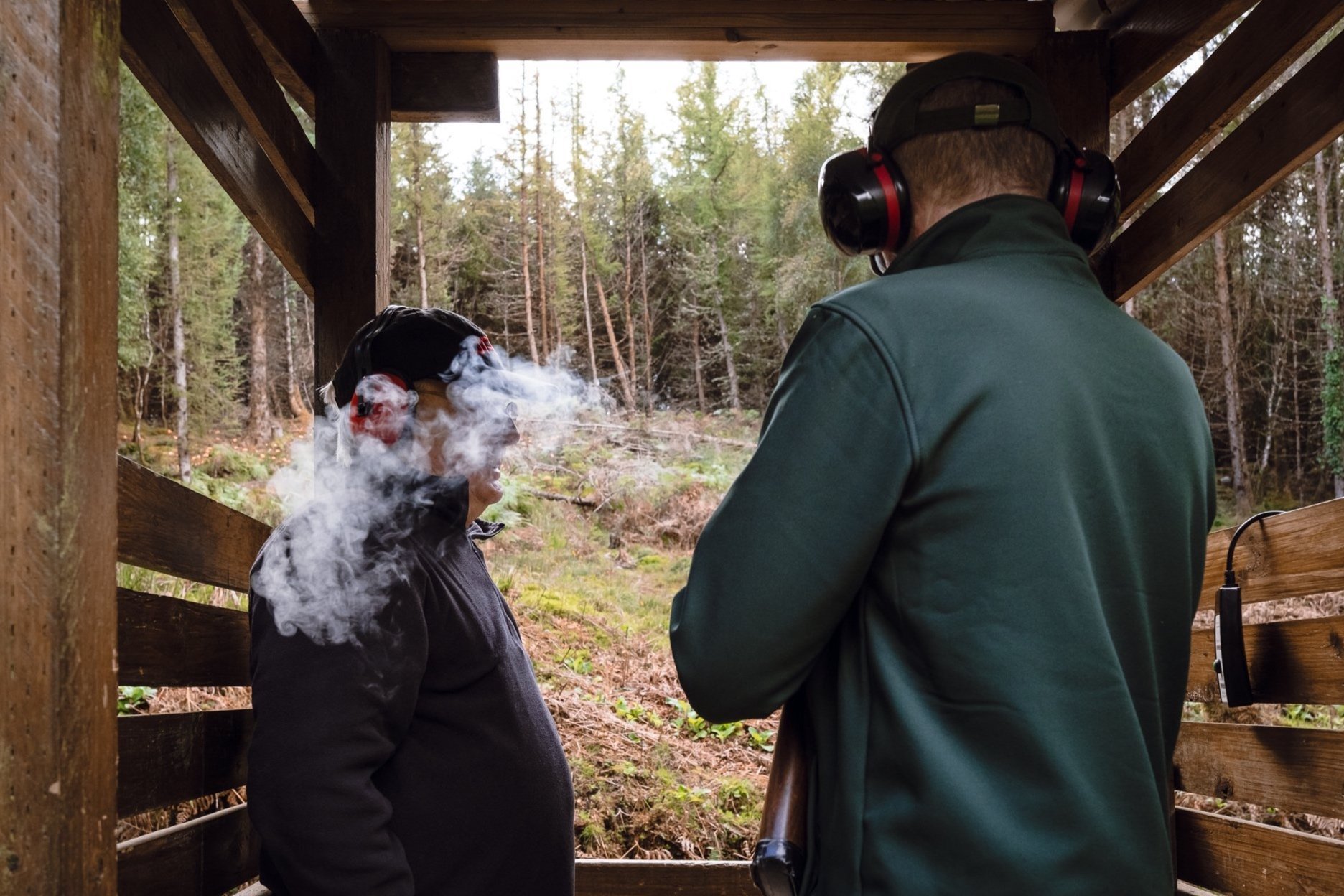 Two men at an outdoor shooting range, one wearing earmuffs and the other smoking while facing each other. The area is wooded with tall trees.