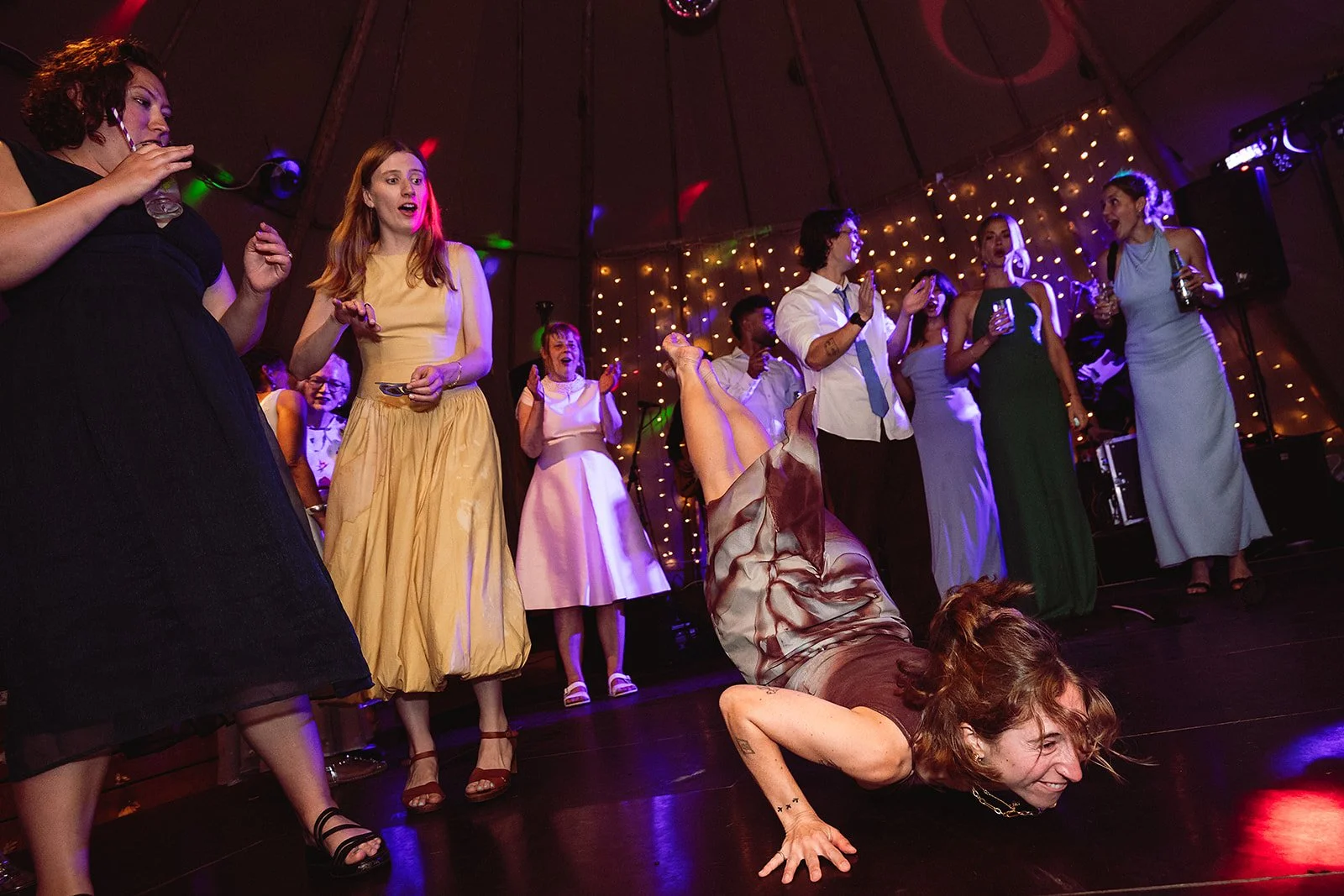 A woman is performing a breakdance move on the floor at a party or celebration, surrounded by onlookers who are smiling and clapping.