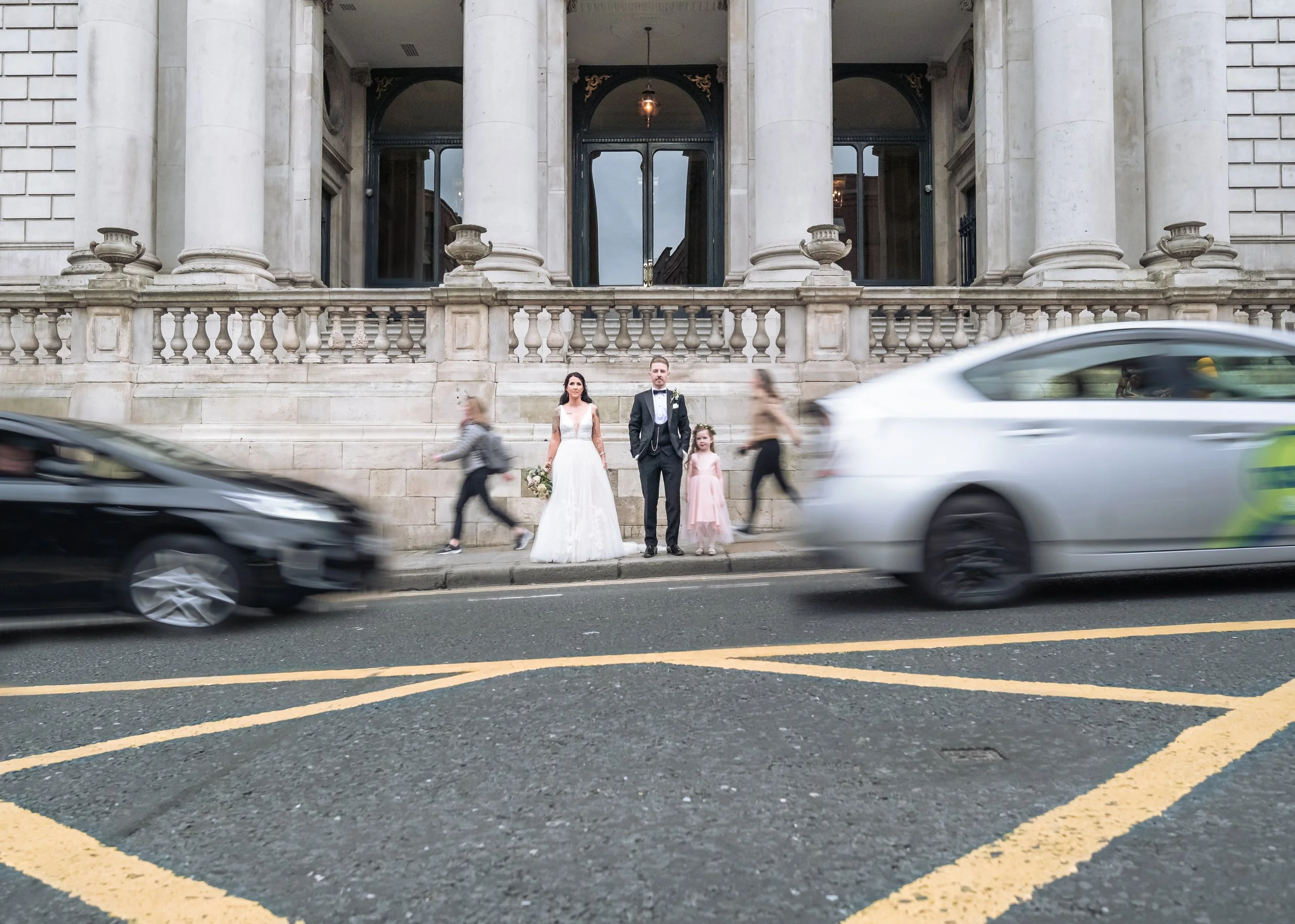 A bride, groom, and flower girl standing on a city sidewalk with blurred cars passing by, in front of a historic building with large columns and decorative urns.