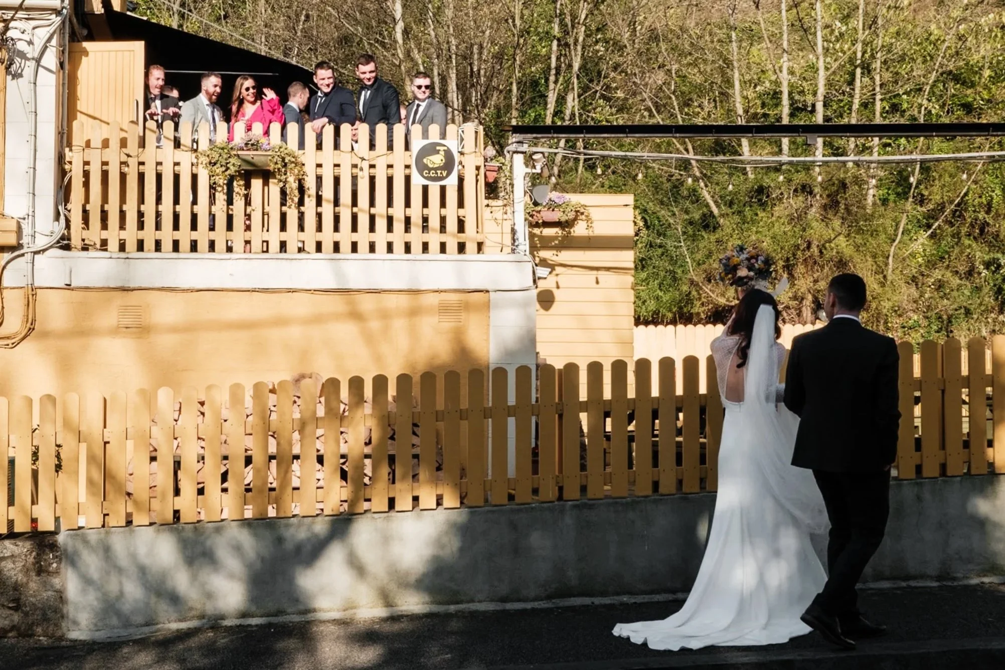 A bride and groom walking outside on a sunny day, with a wooden fence separating them from a group of people on a balcony above. The group on the balcony is smiling and waving, possibly watching the couple.