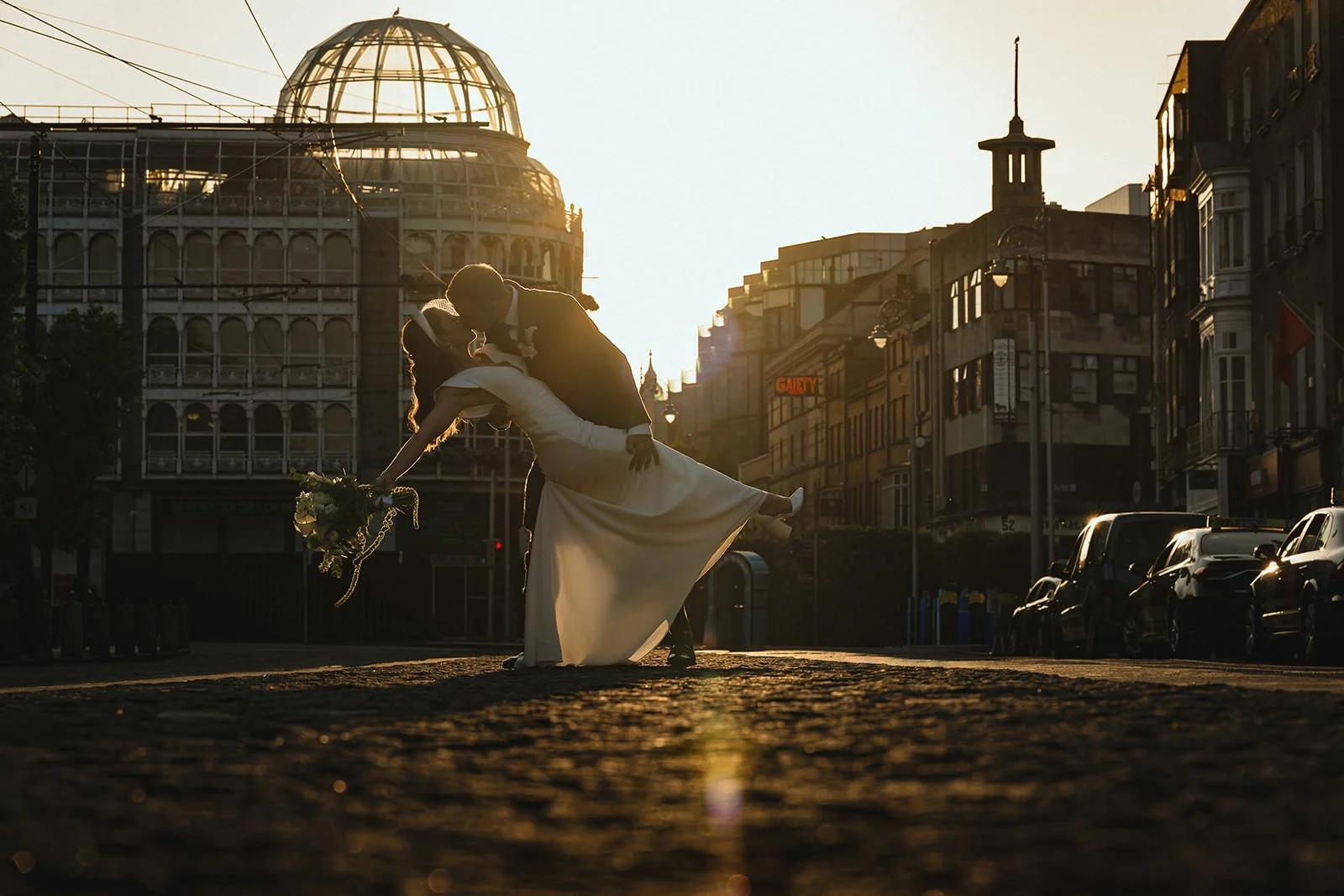 A bride and groom sharing a dance in a city street at sunset, with the groom dipping the bride backward while holding her bouquet, and city buildings in the background.